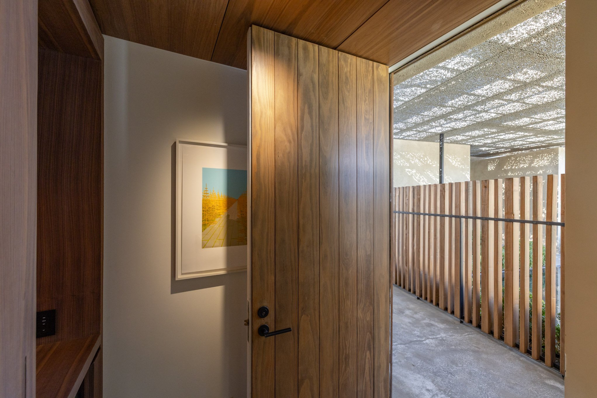 A wooden door is open, revealing a small porch area with wooden fence panels and a ceiling with a perforated metal or mesh design, casting patterned shadows on the outdoor surfaces.