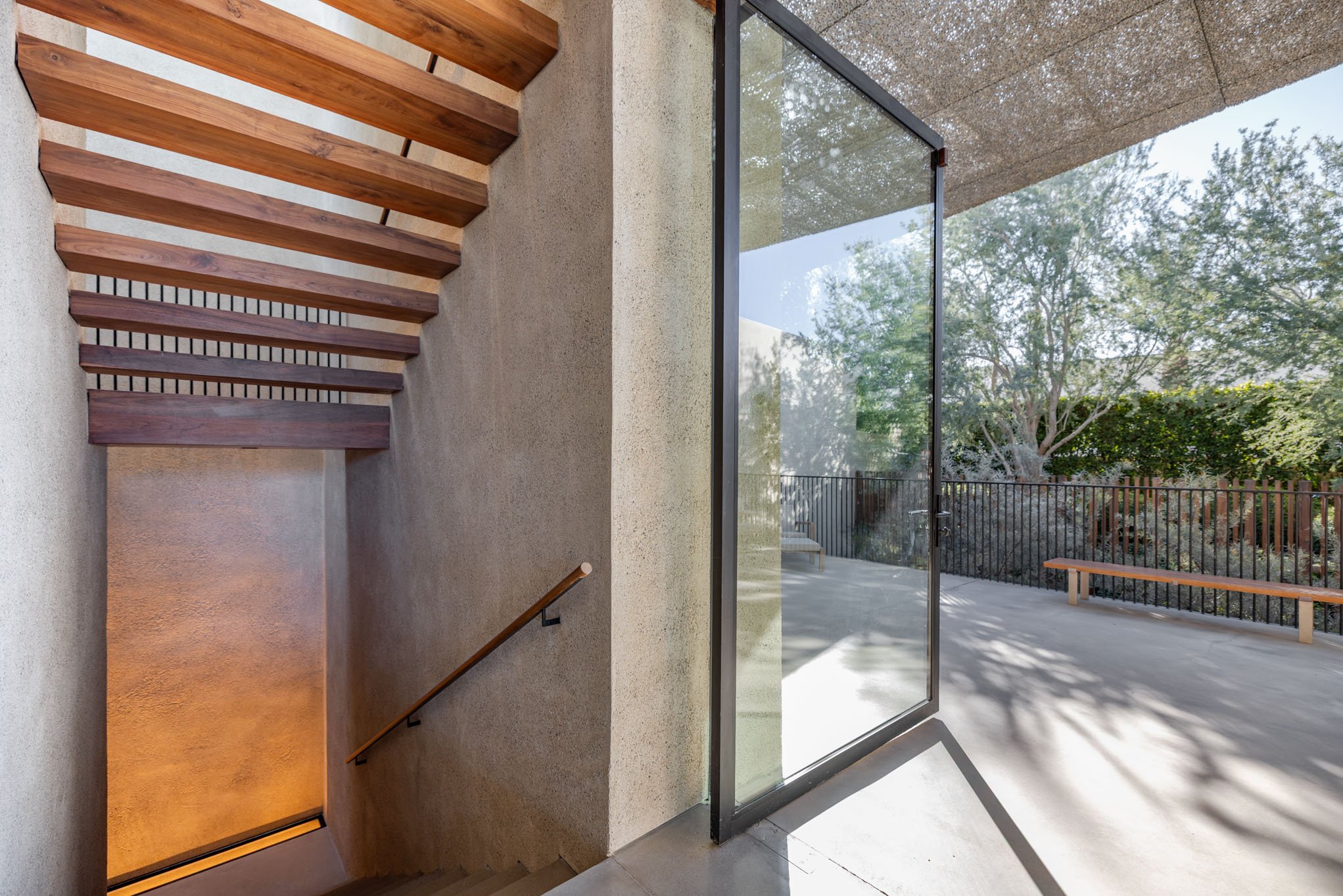 A modern outdoor patio with a concrete floor, a glass door, a wooden bench, trees and bushes in the background, and a stairway with wooden steps leading downward.