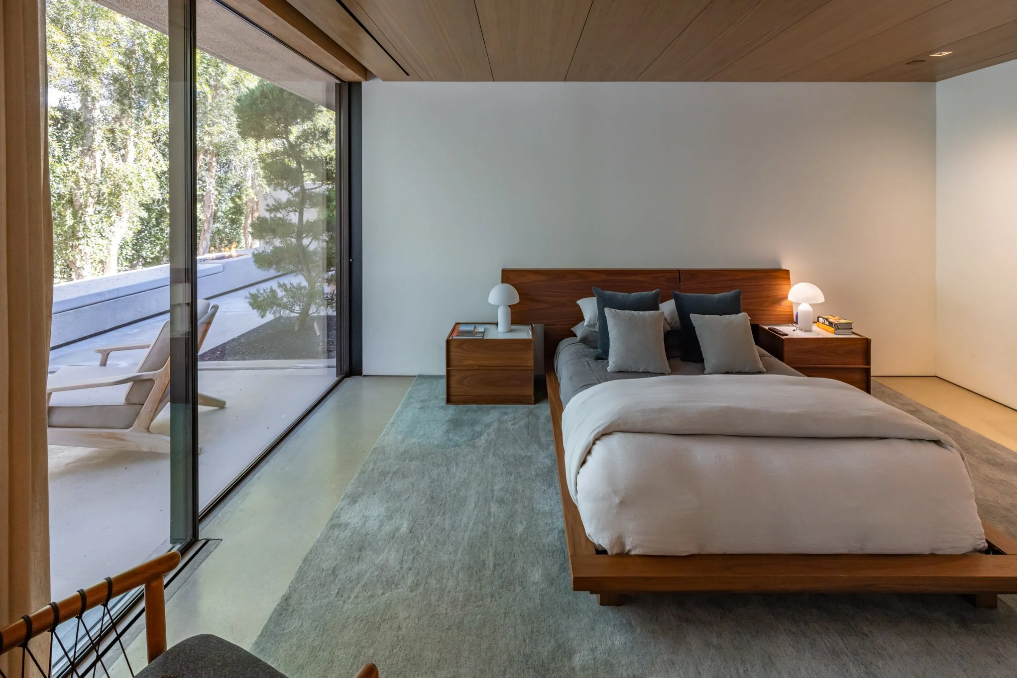 Modern bedroom with a large bed, multiple pillows, nightstands with lamps, sliding glass door leading to a balcony with outdoor seating, and a view of trees outside.