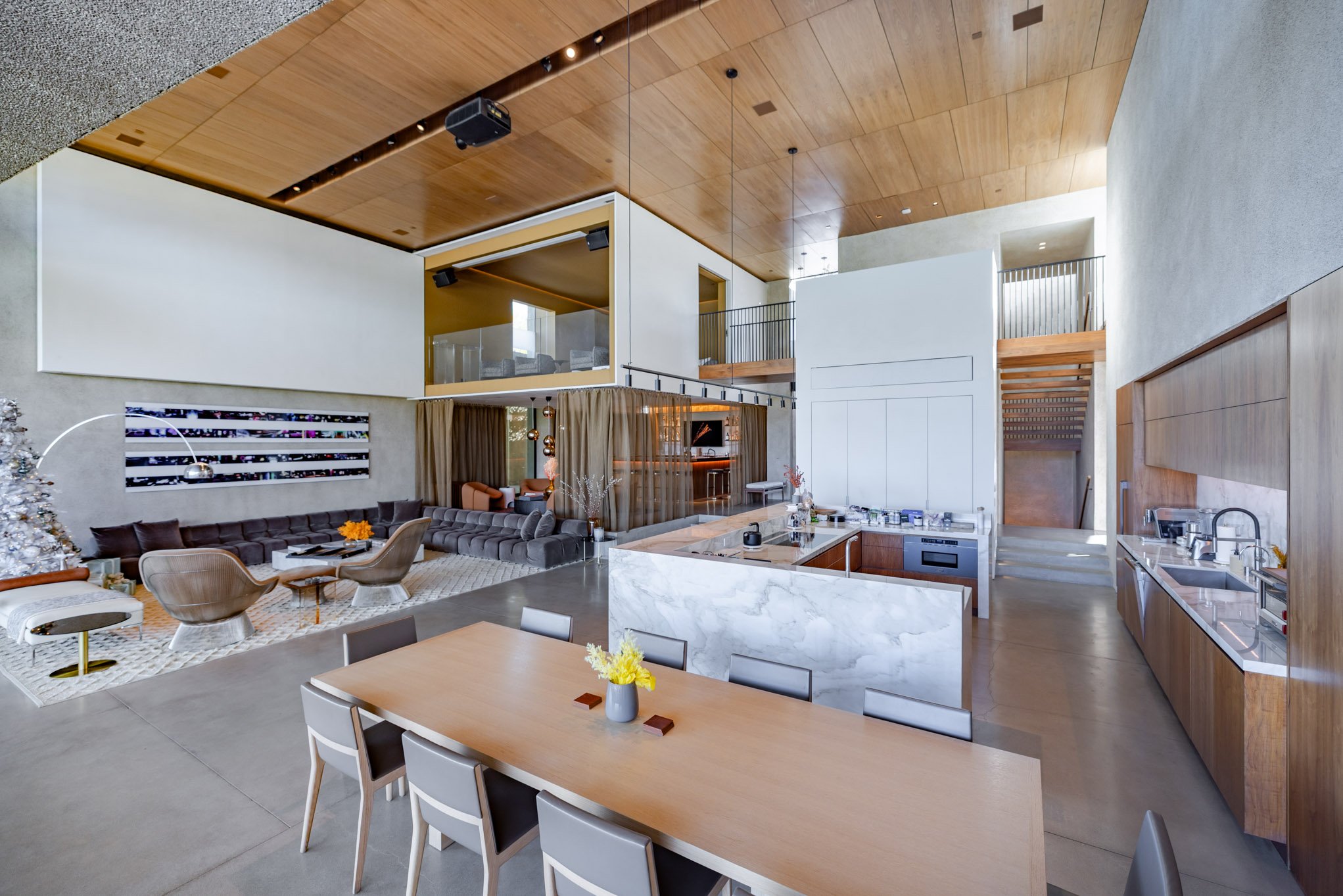 Open-concept modern living room and kitchen with high ceilings, wooden and white walls, a wooden ceiling, a large dining table, a marble kitchen island, and a cozy seating area with a sofa and chairs.