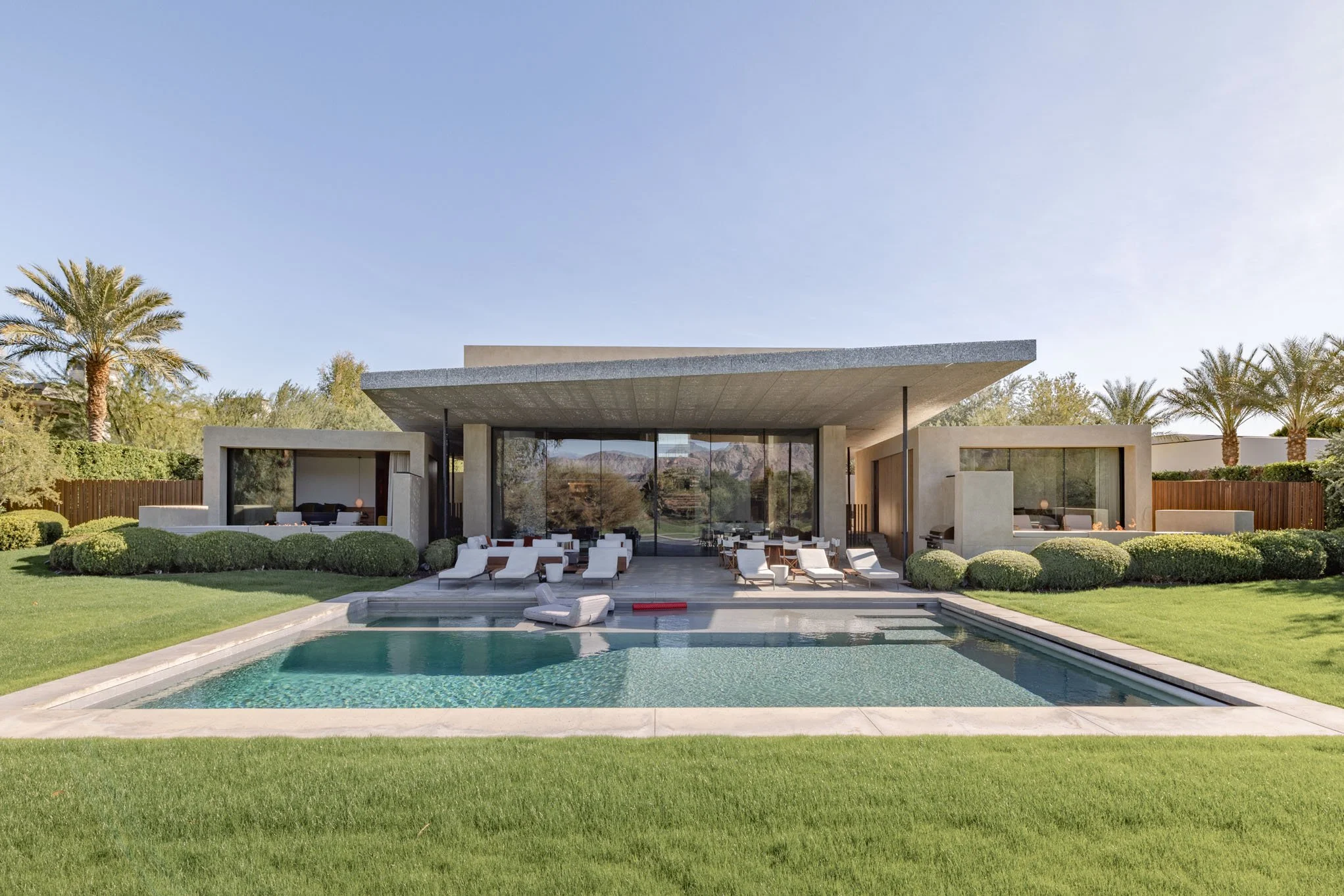 Modern house with large sliding glass doors, surrounded by well-manicured lawn, palm trees, and shrubs, featuring an outdoor swimming pool with lounge chairs and seating area.