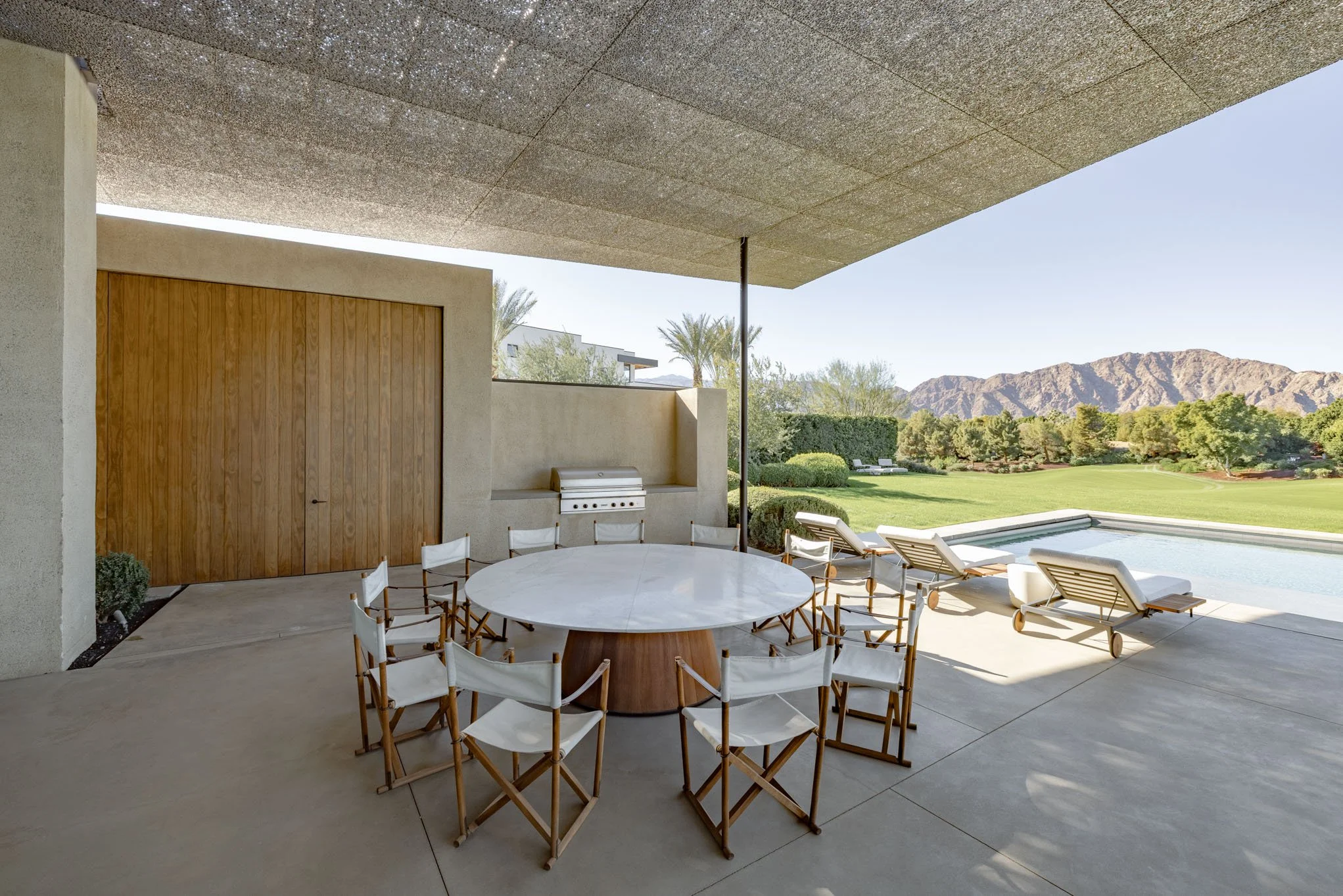 Outdoor patio with a round dining table surrounded by white and wood chairs, adjacent to a swimming pool and lounge chairs overlooking a green landscape with mountains in the background.