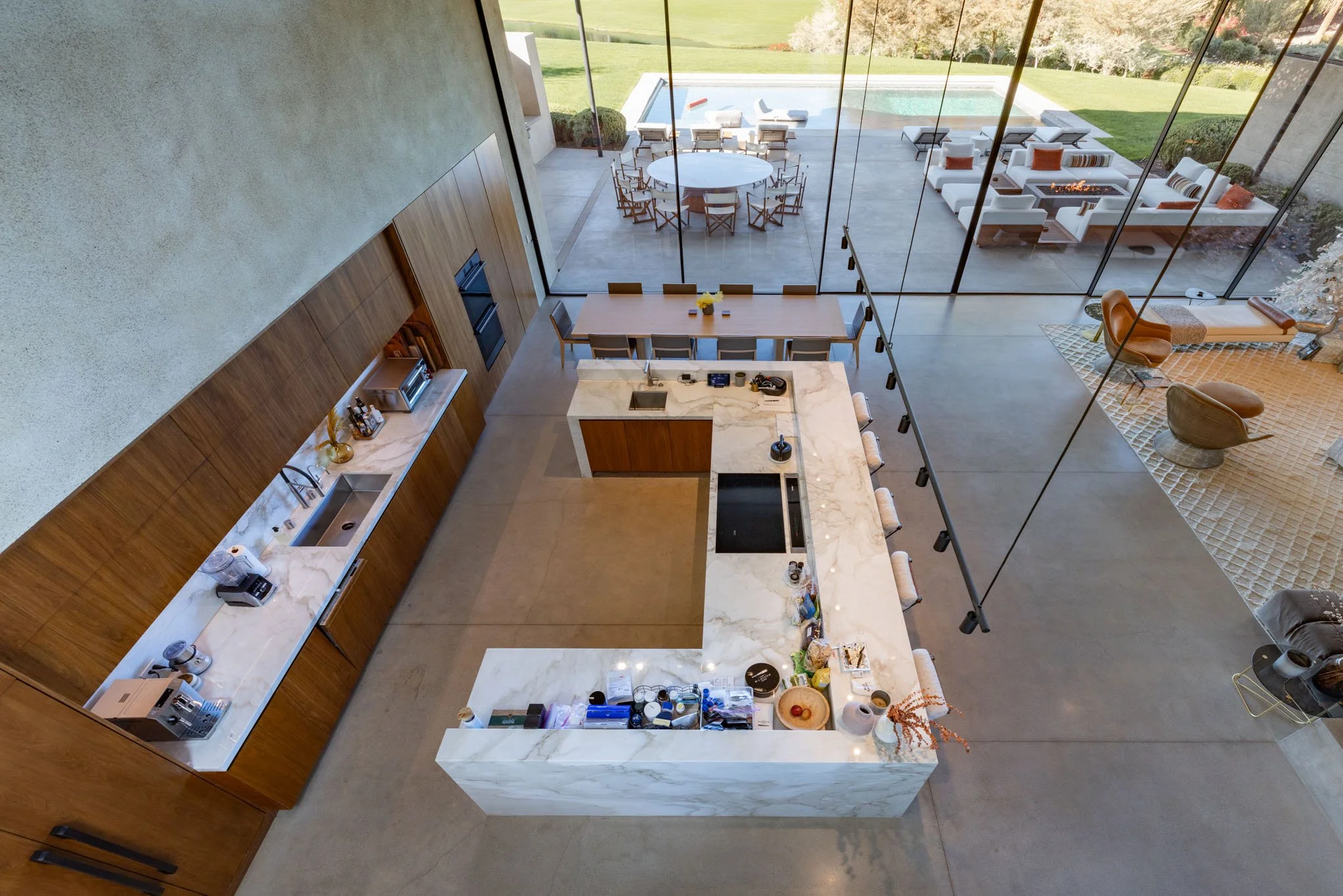 Interior view of a modern kitchen with a large marble island counter, wooden cabinets, and open access to an outdoor patio with seating areas and a swimming pool.