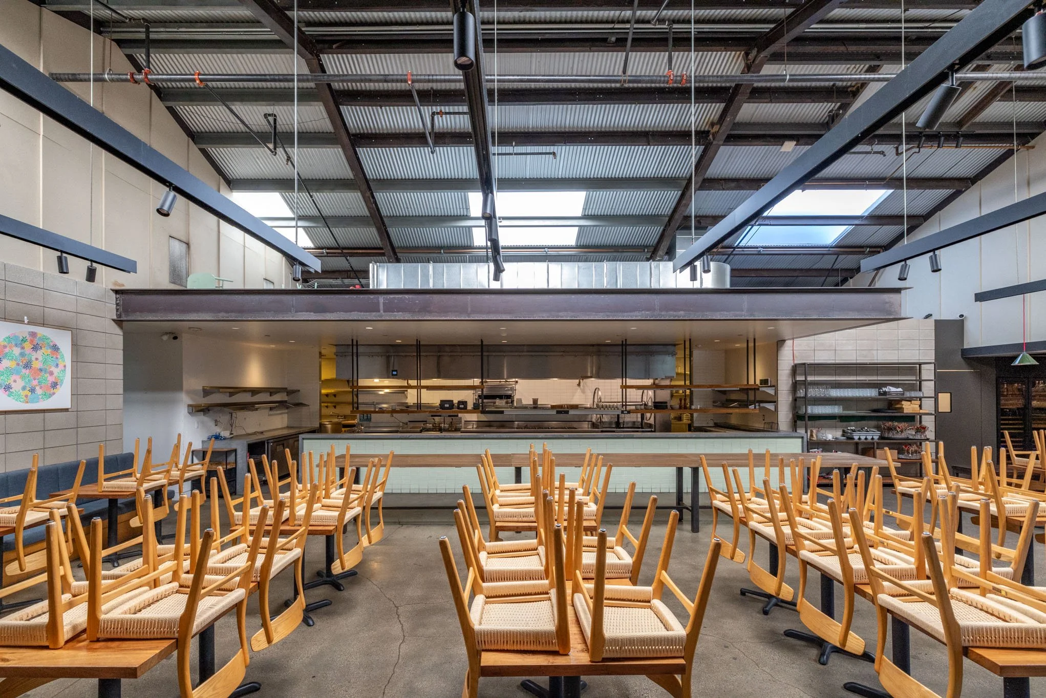 Empty dining area with stacked chairs and an open kitchen under a metal roof in a restaurant or cafe.