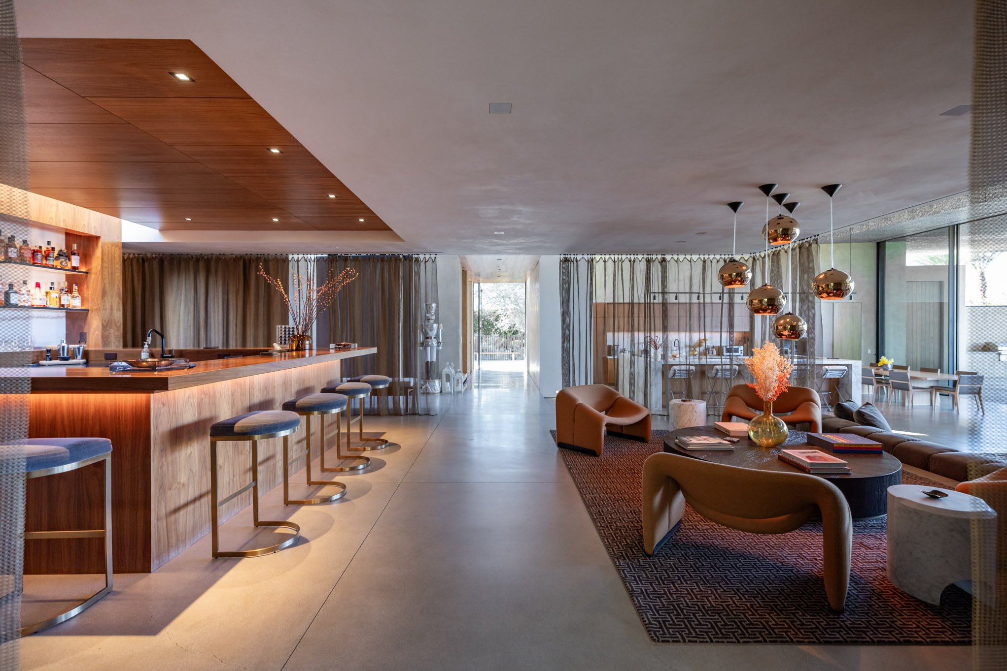 Modern hotel lobby with a bar area featuring four bar stools, a wooden counter, and a backlit shelf with liquor bottles. The seating area includes contemporary chairs, a round coffee table with magazines, and decorative vases with flowers. Large wind