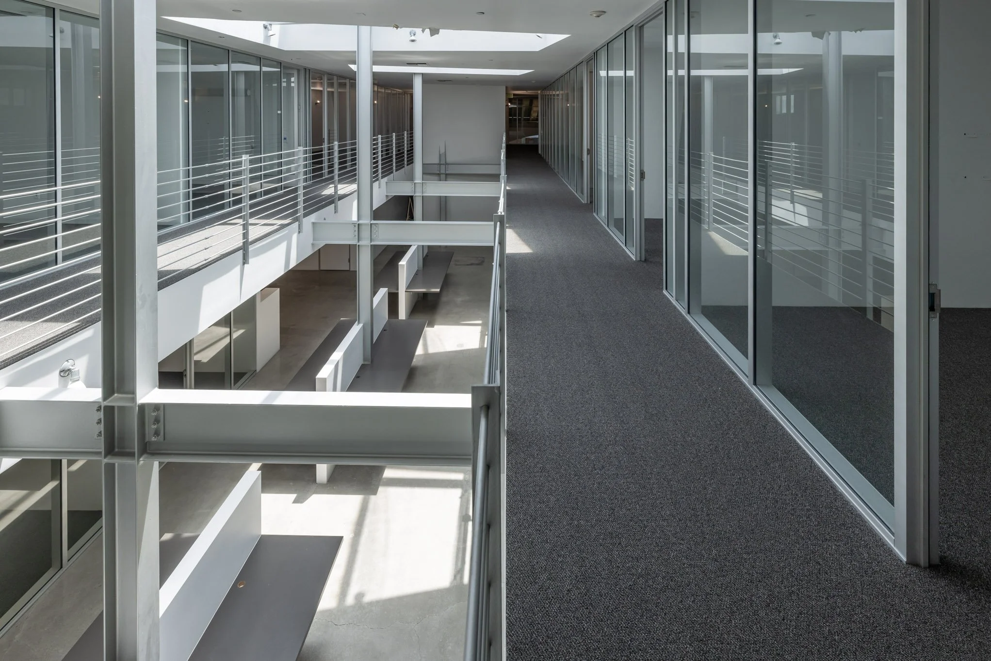 Modern office interior with glass walls, gray carpeted hallway, and railing overlooking the lower level.