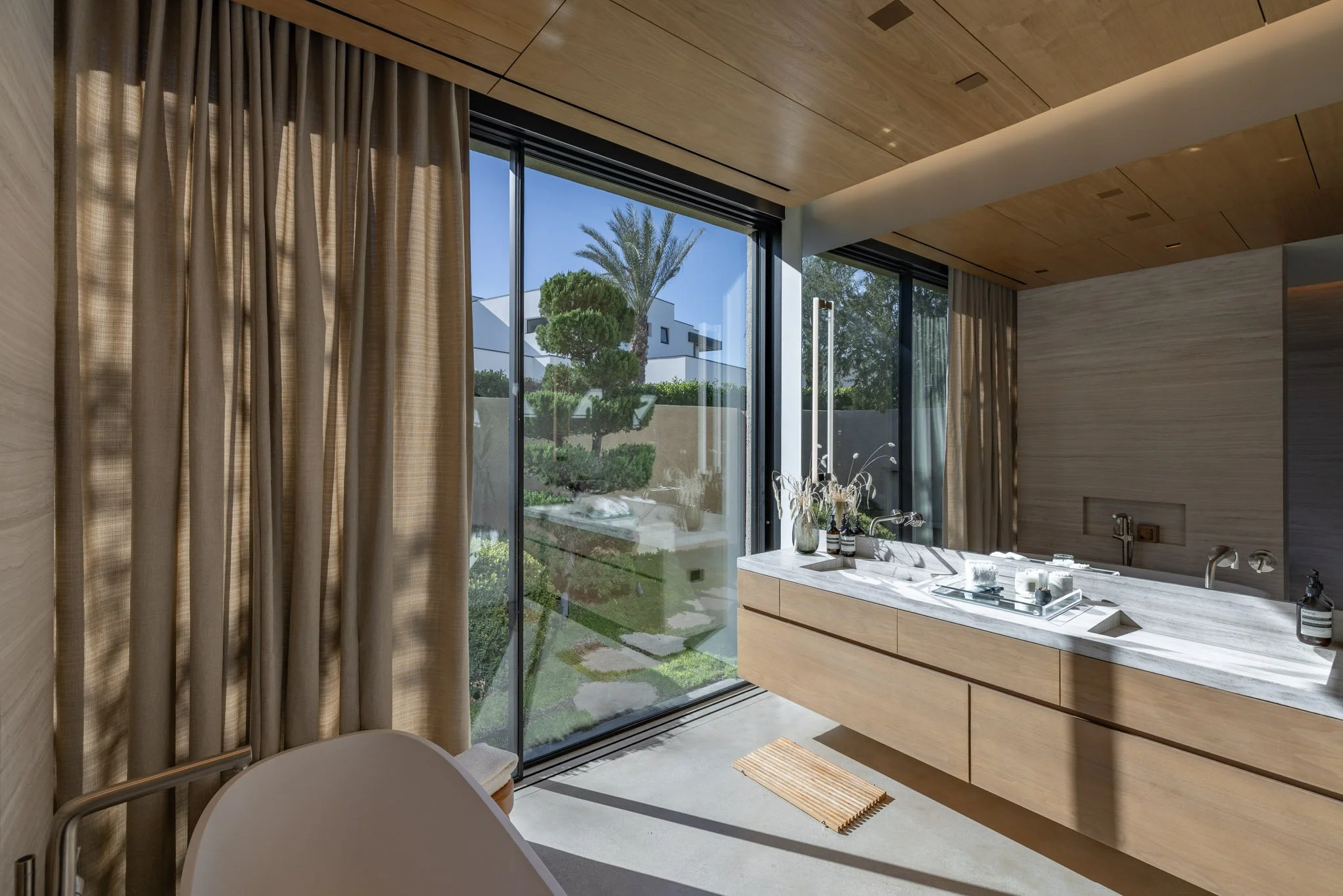 Modern bathroom with large glass windows, beige curtains, wooden ceiling, and a marble sink countertop with decor