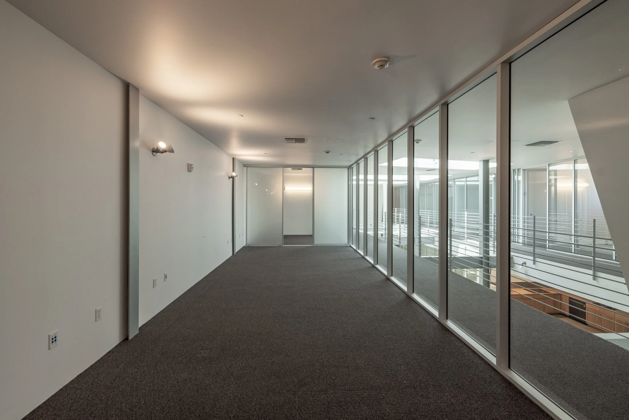 Empty indoor corridor with glass windows on the right, white walls, and ceiling lights.