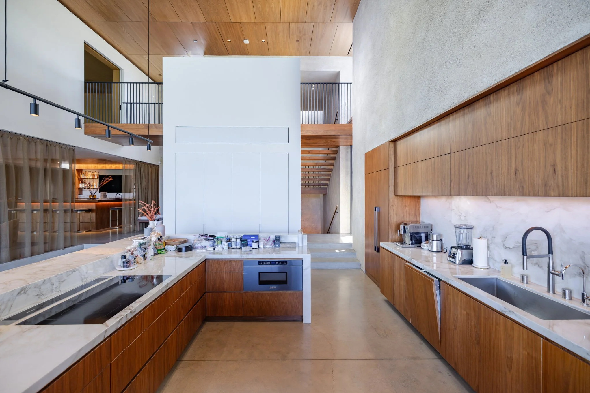 Modern kitchen with wooden cabinets, marble countertops, and a built-in oven, featuring a double sink and various appliances, with a staircase and upper level visible in the background.