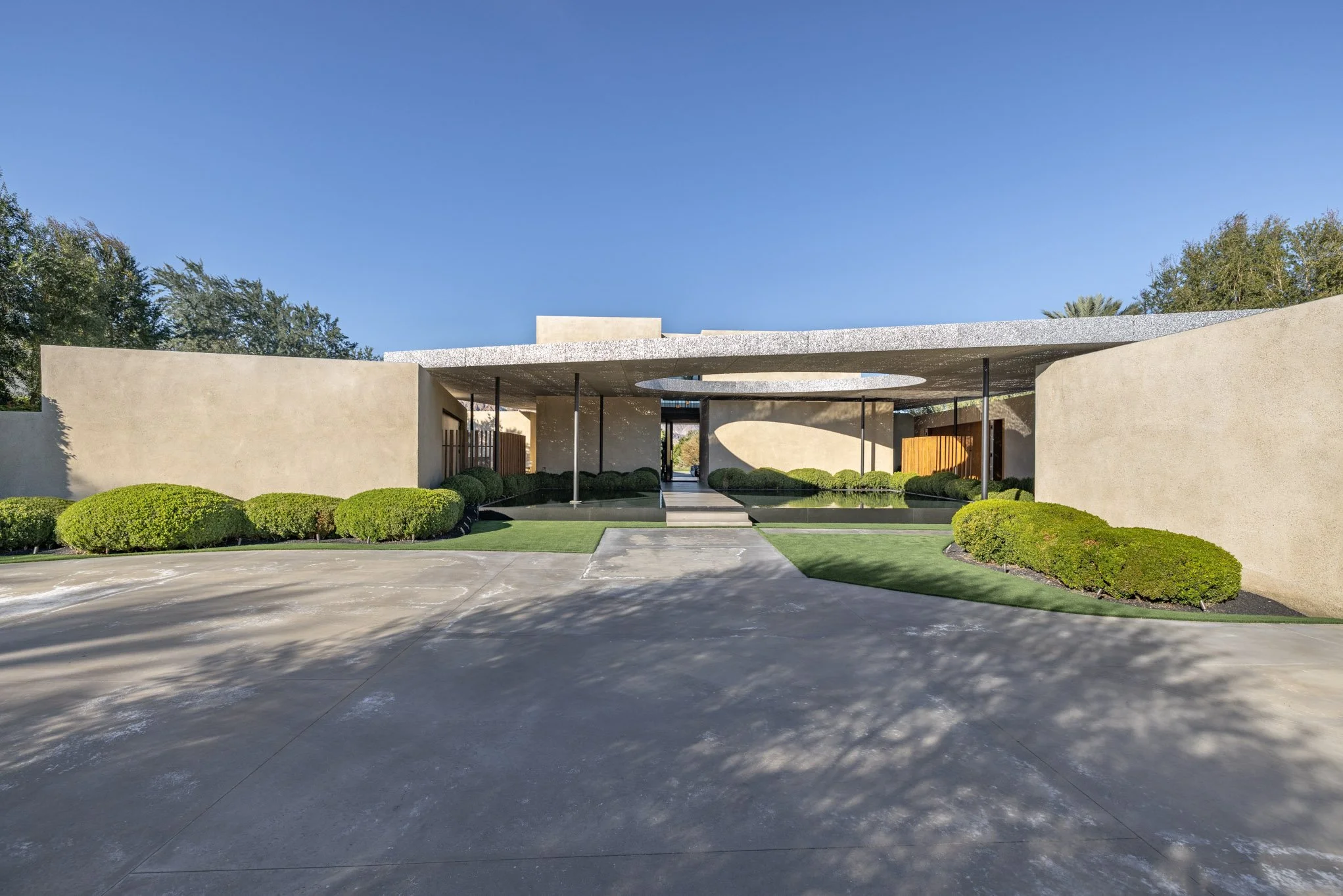 Modern single-story building with beige stucco walls, a flat roof with a circular cutout, surrounded by green bushes and a manicured lawn, with a concrete driveway in front, under a clear blue sky.