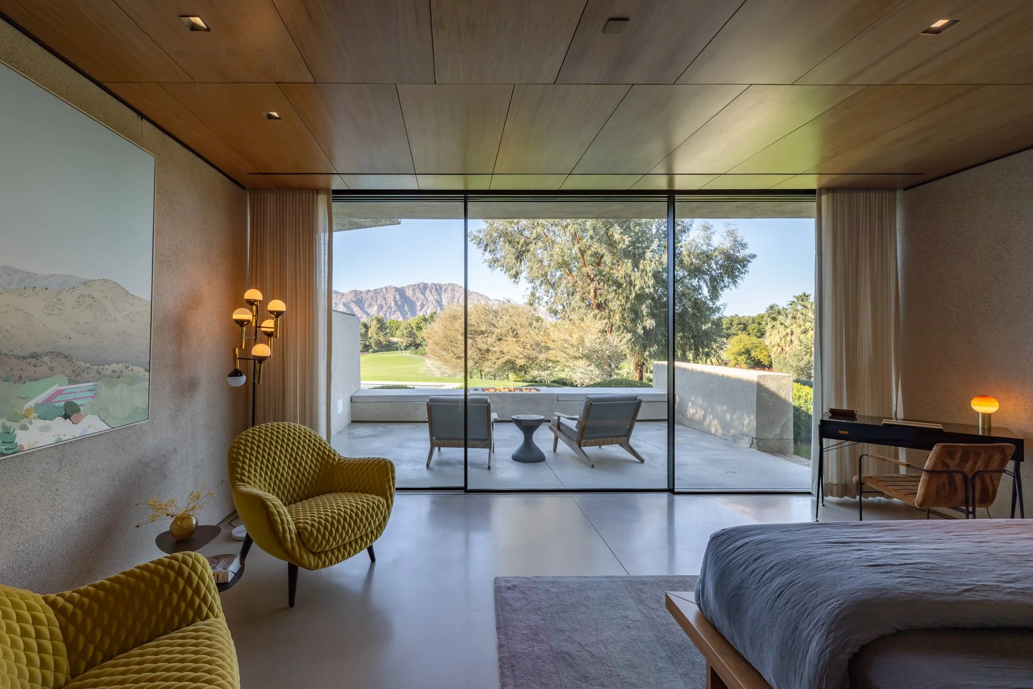 Modern bedroom with large glass sliding doors leading to an outdoor patio with chairs and a scenic mountain view.