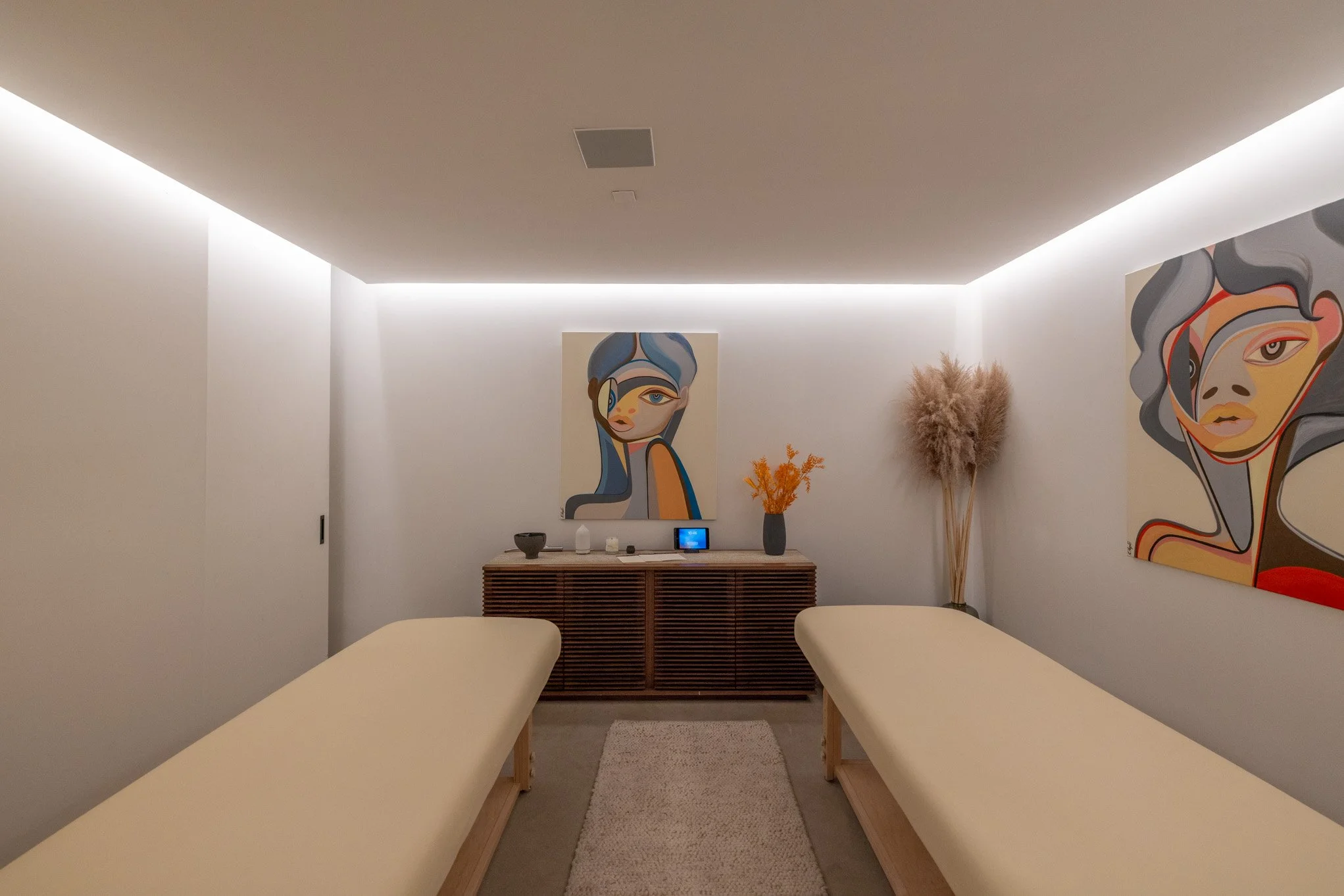 A modern massage or relaxation room with two massage tables, abstract art paintings of women's faces on the walls, a wooden cabinet with decorative objects, and dried plants in vases.