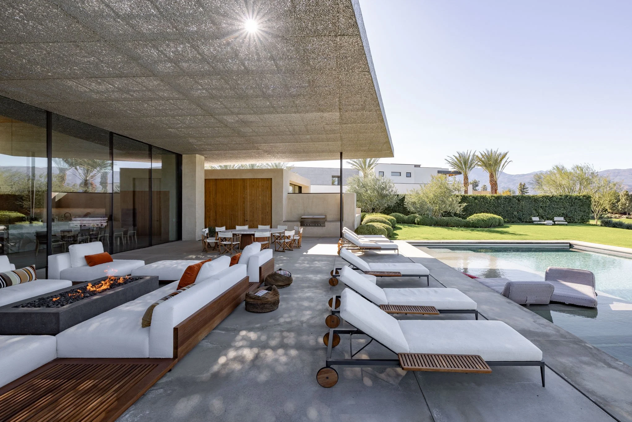 Modern outdoor patio with white cushioned lounge chairs, a rectangular fire pit, a dining table with chairs, and a swimming pool surrounded by green grass, palm trees, and a clear sky.