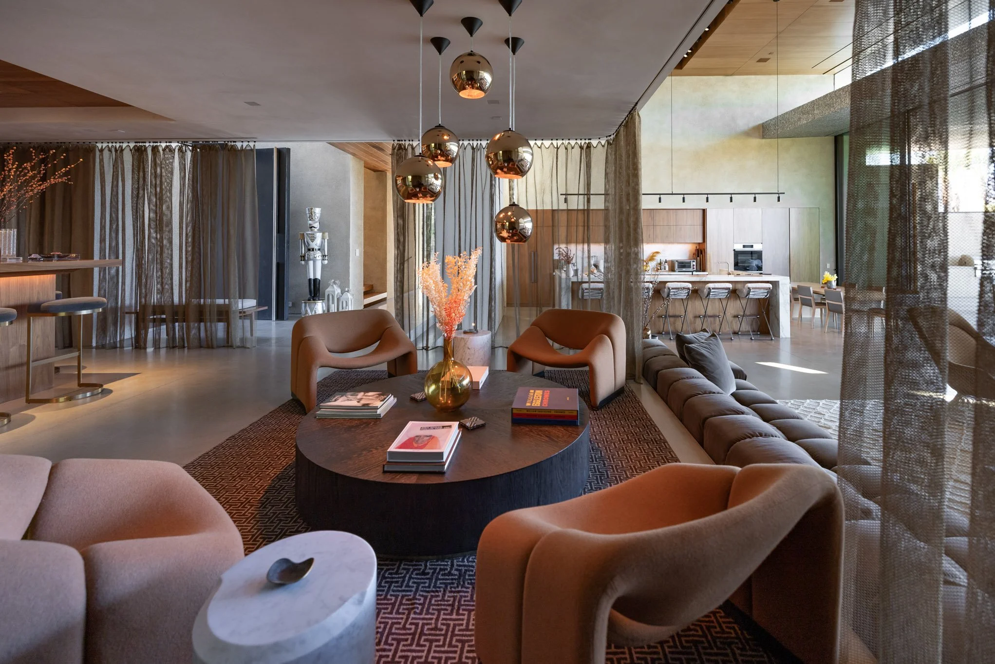 Modern open-concept living room with a round coffee table, beige and brown chairs, a long sectional sofa, hanging pendant lights, and view into a kitchen with bar stools and dining area.