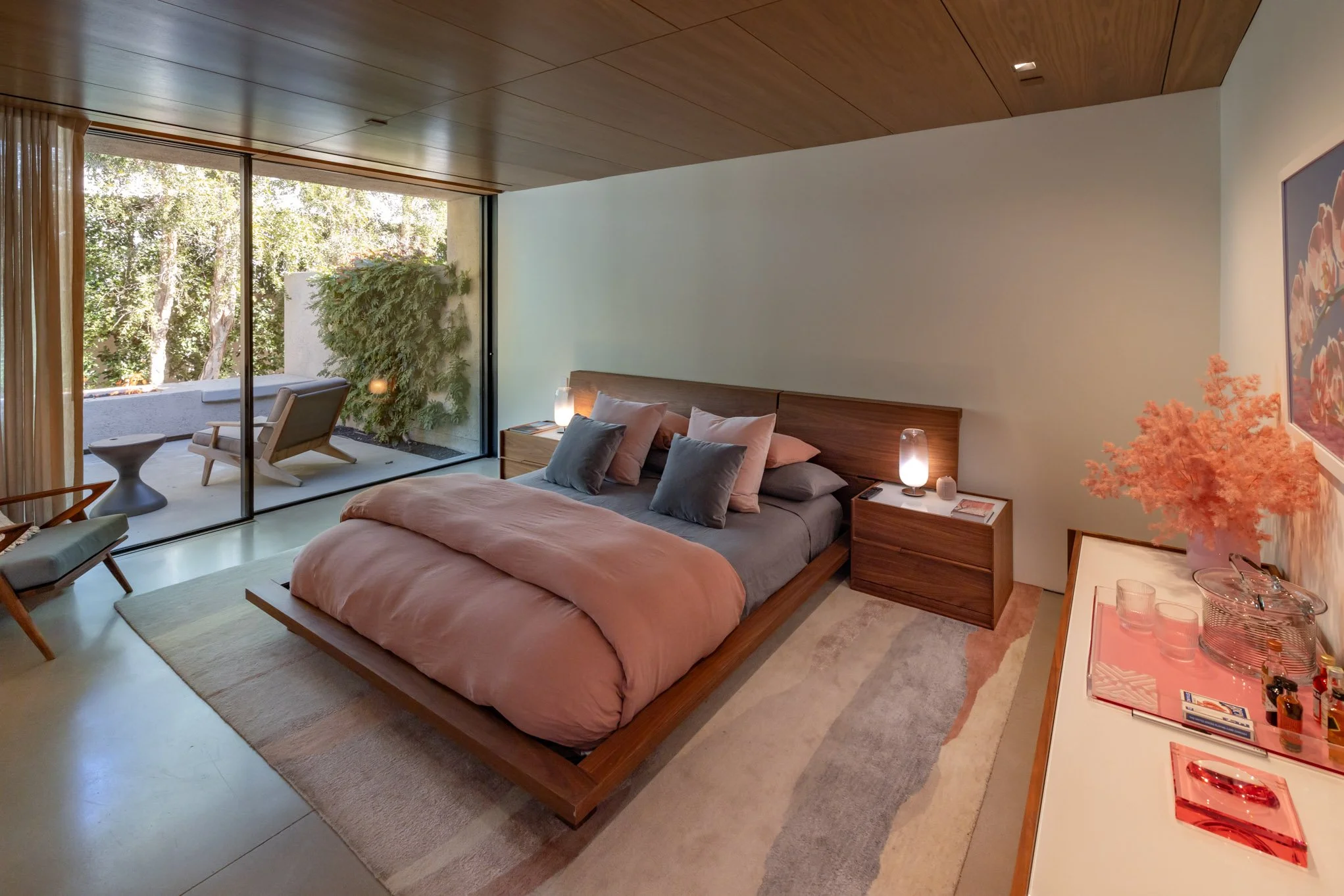 Modern bedroom with a large bed, gray and pink bedding, and several pillows. A sliding glass door leads to an outdoor patio with a chair and small table. Nightstands with lamps are on either side of the bed. A buffet with pink decor and artwork adorn