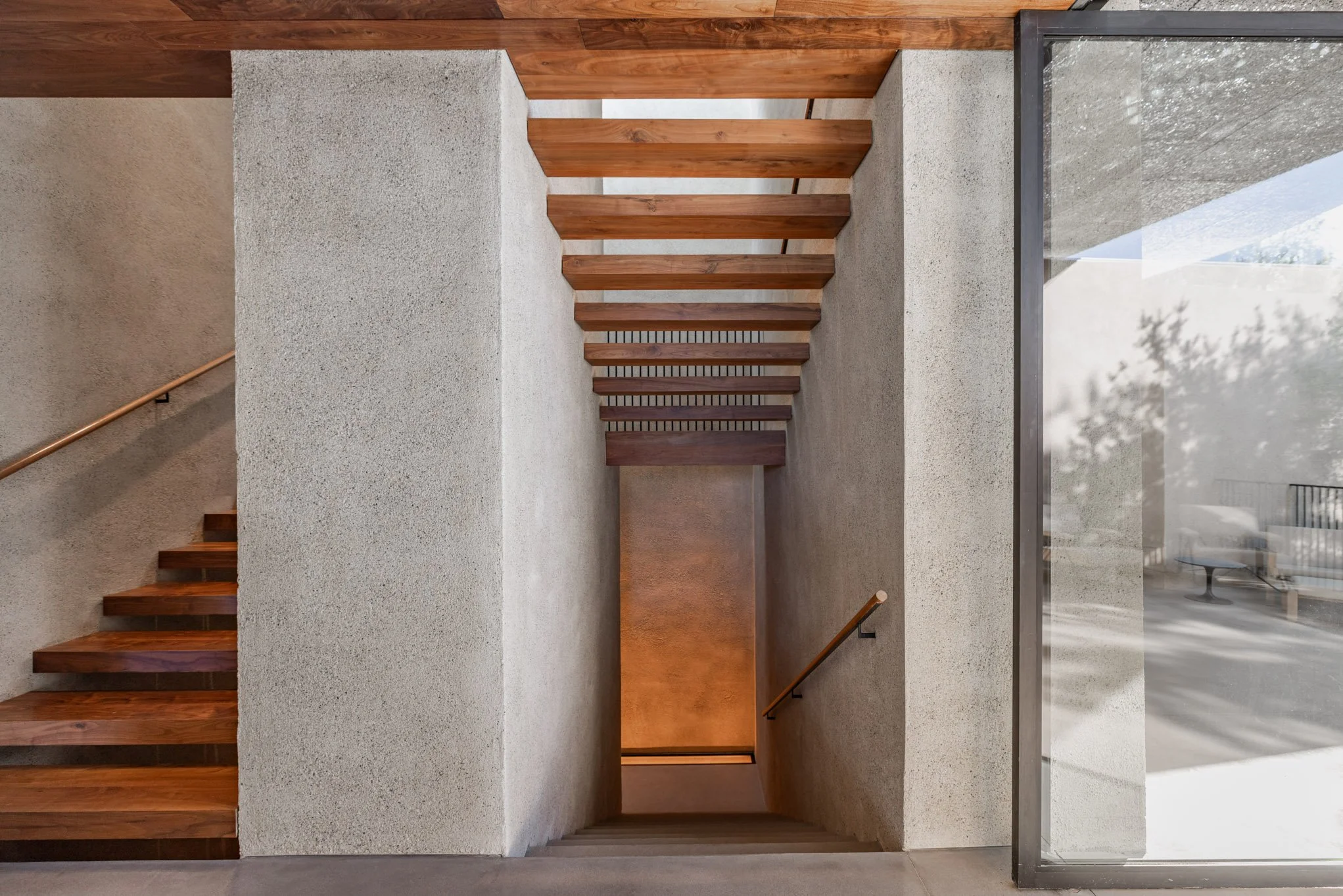 Interior view of a staircase with wooden steps and handrails, textured concrete walls, and a large front glass window showing outdoor furniture.