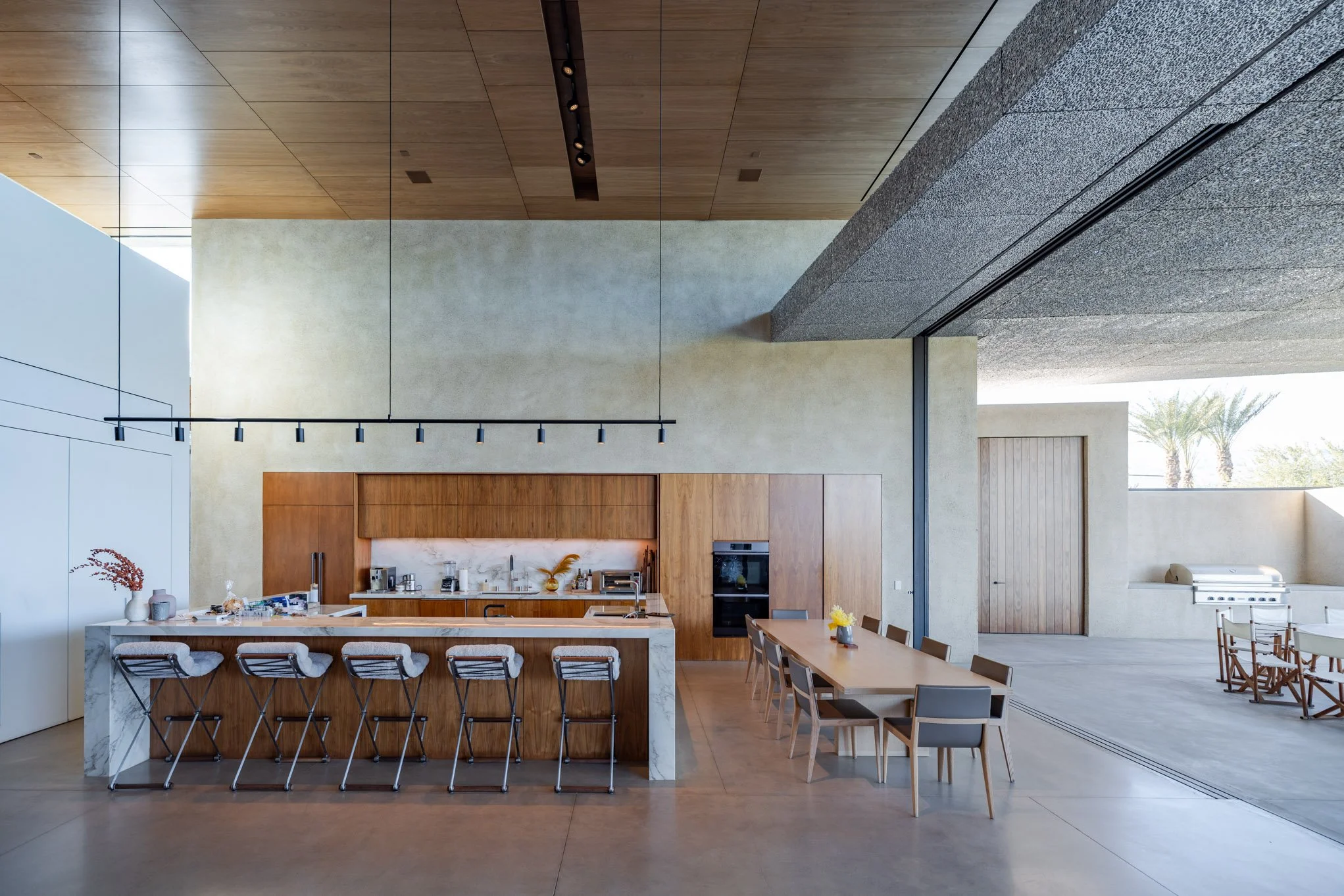 Modern open-concept kitchen and dining area with wooden accents, marble island, bar stools, and a large table, featuring large windows and palm trees outside.