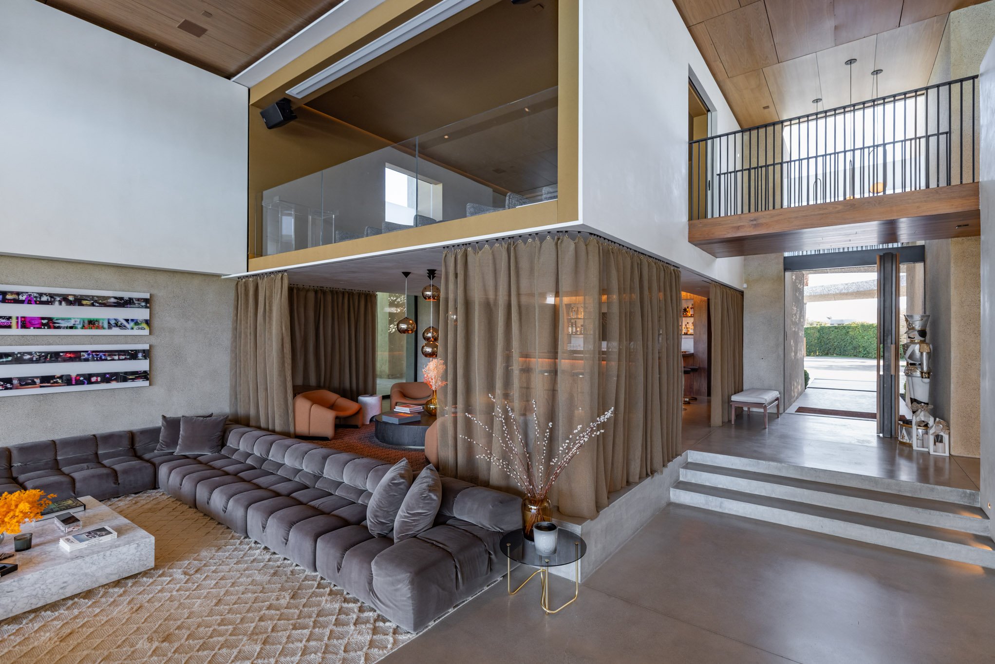 Modern living room with a large gray sectional sofa, beige curtains around a seating area, a coffee table with books, artwork on the wall, and a staircase leading to an upper level. There is also a small round side table with a vase and decor, and a 