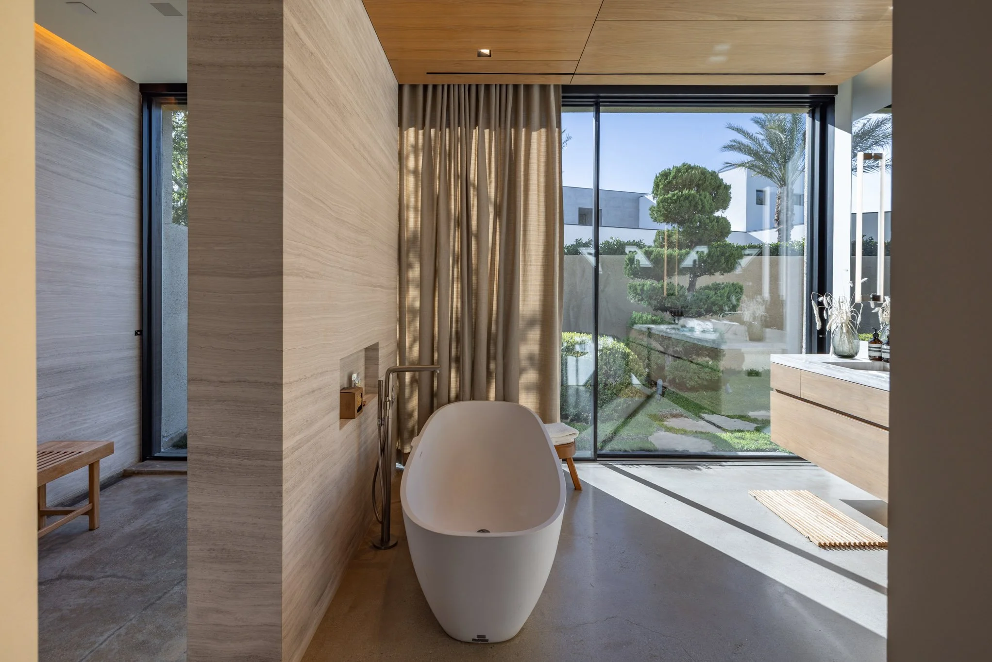 Modern bathroom with a freestanding bathtub, large floor-to-ceiling window with beige curtains, and view of a landscaped garden with trees outside.
