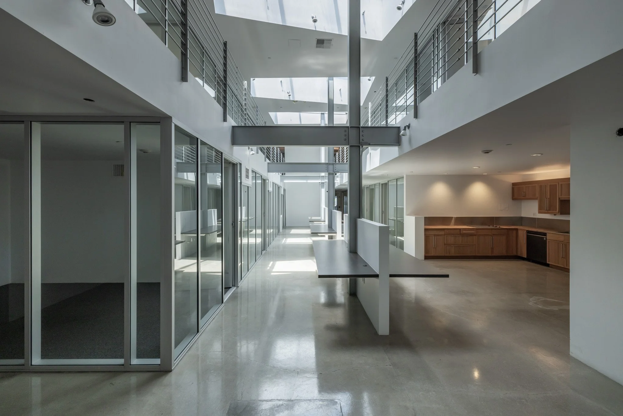 Empty modern office space with glass-walled conference rooms on the left, a countertop kitchen area with wooden cabinets on the right, and a staircase with metal railings on the upper level, with daylight streaming in from skylights.