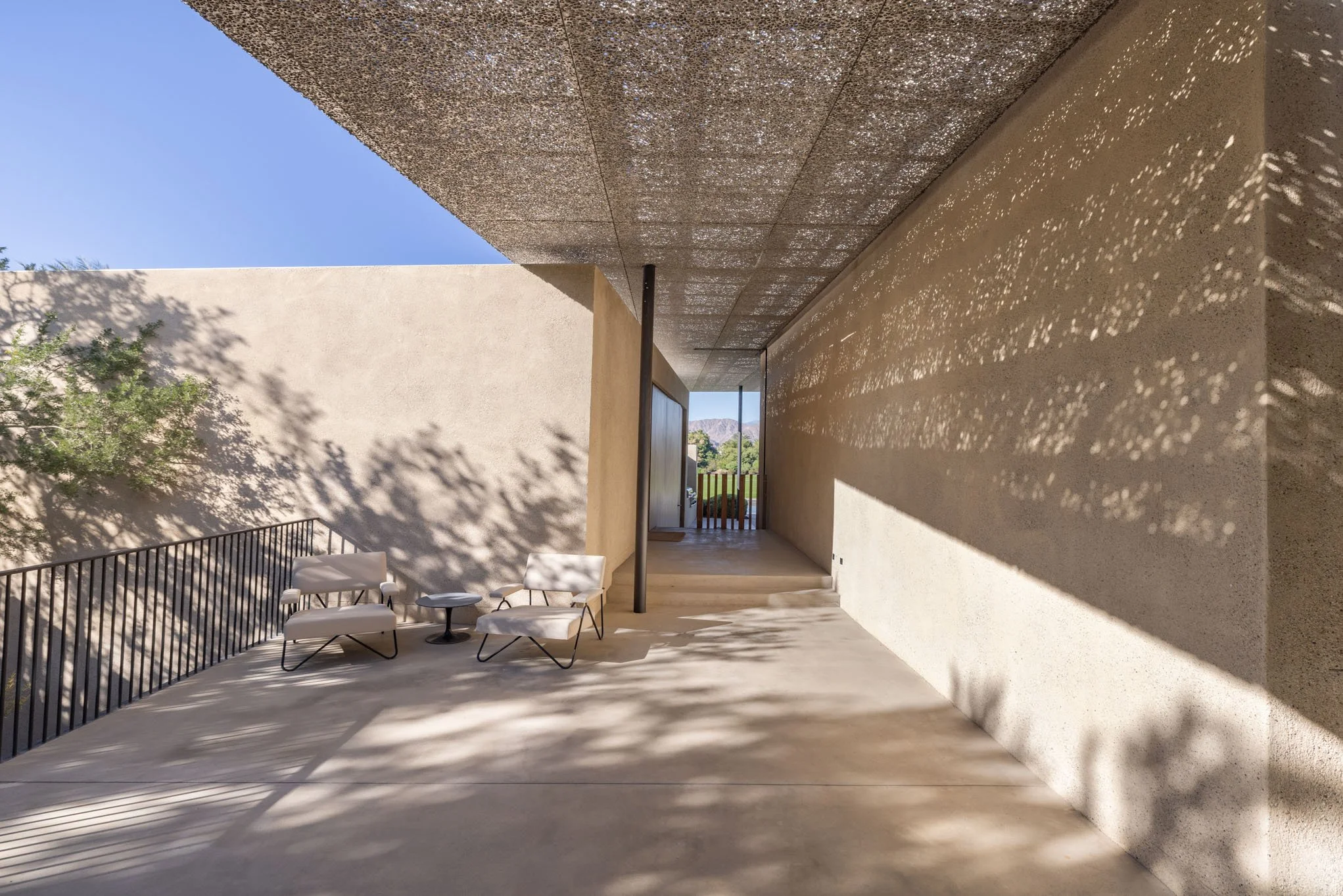 Modern outdoor balcony with two white chairs, a small side table, and black metal railing, with shadows of leaves cast on the wall and floor, and a view of green hills and mountains in the distance.