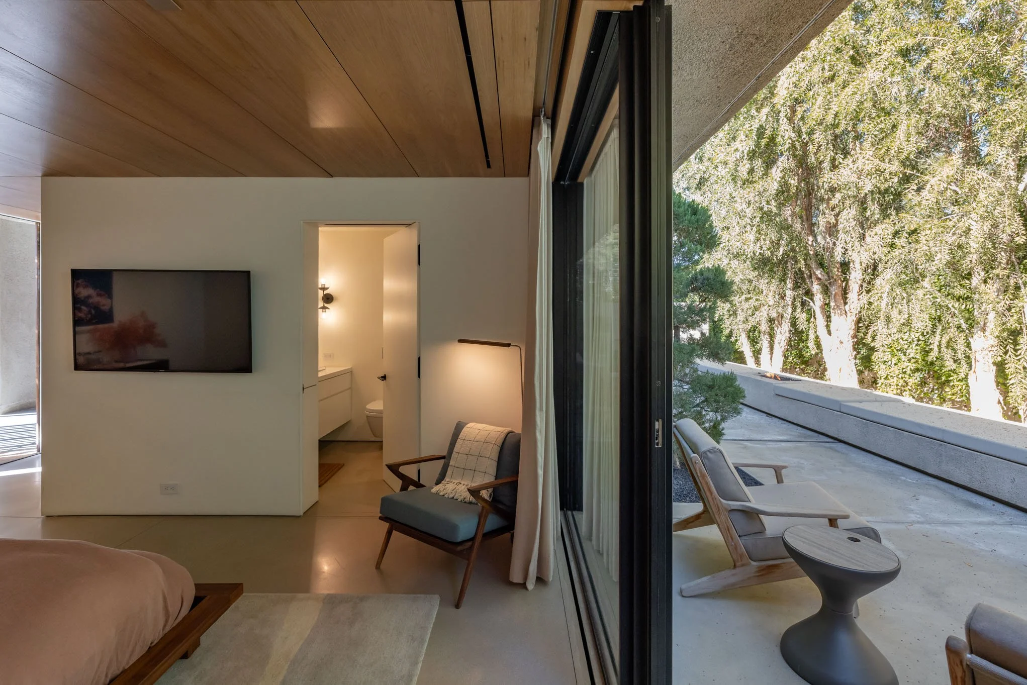 Modern bedroom with a large sliding glass door leading to a balcony with outdoor chairs and trees outside.