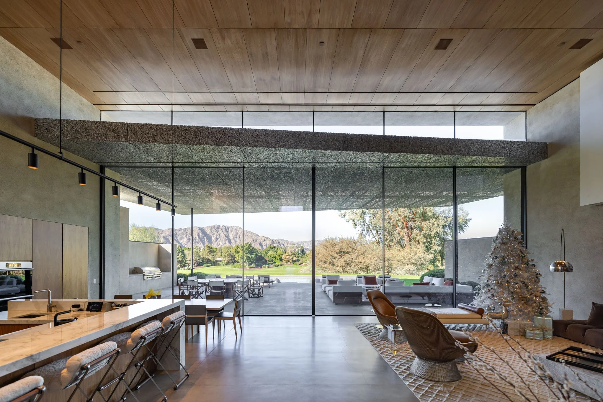 Modern open-concept living space with large glass windows overlooking a green landscape and mountains, decorated with a Christmas tree, lounge chairs, and contemporary furniture.