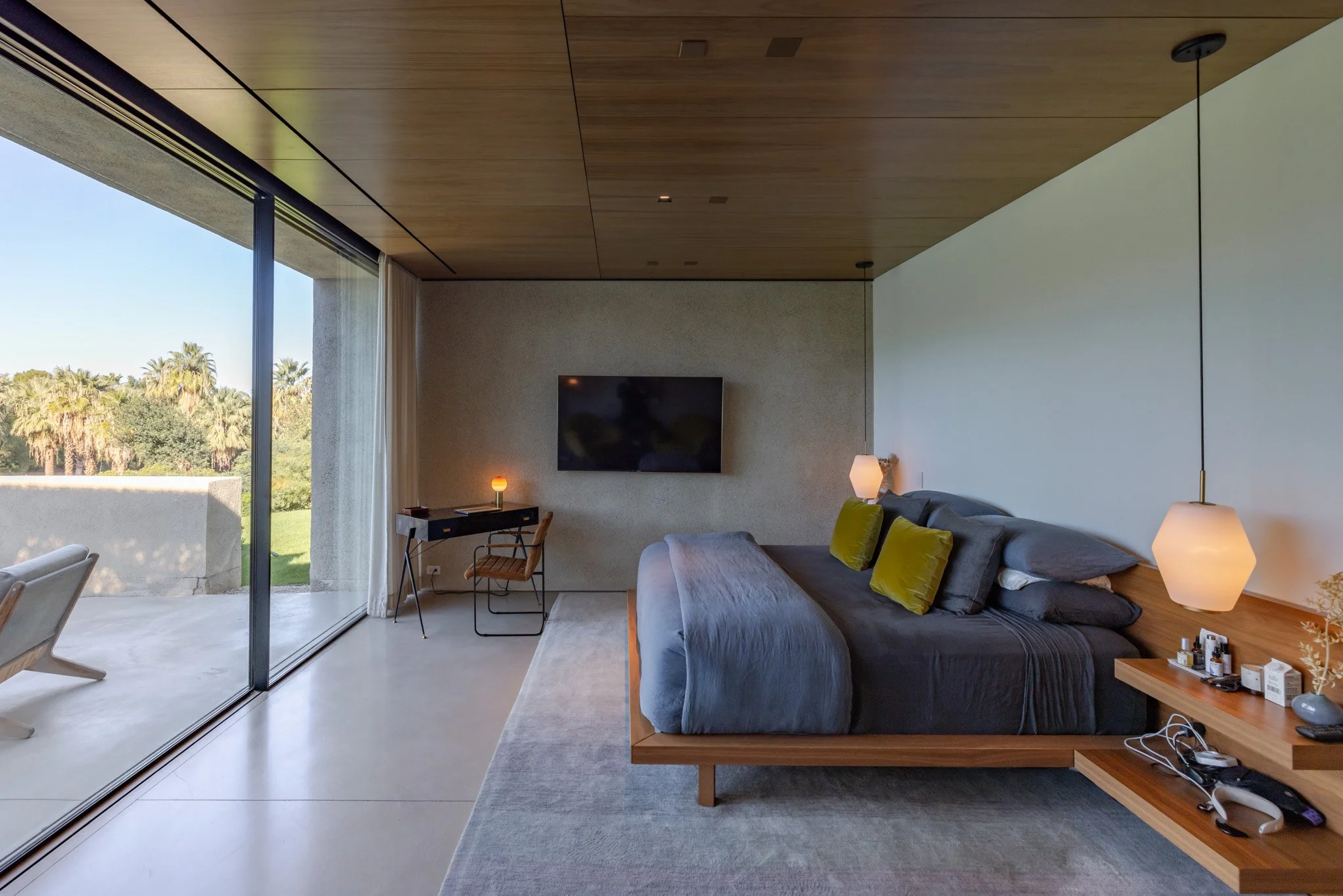 Modern bedroom with large sliding glass doors that open to a patio with greenery. The room features a bed with gray linens, green pillows, a floating wooden bedside table, pendant lights, a flat-screen TV on the wall, and a small desk with a chair an