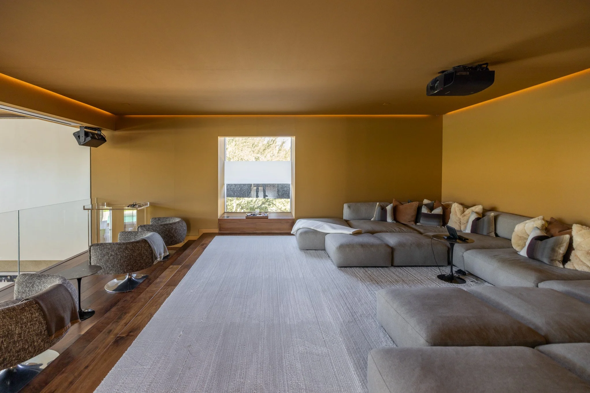 Modern living room with gray sectional sofa, assorted pillows, small side table with phone, wooden floor, large window with outside view, beige walls, recessed ceiling lighting, and ceiling-mounted speakers.