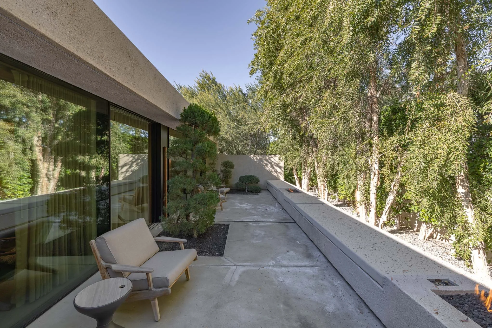 Modern outdoor patio with beige chairs, small round side table, concrete floor, tall trees, and a fire feature in the corner.