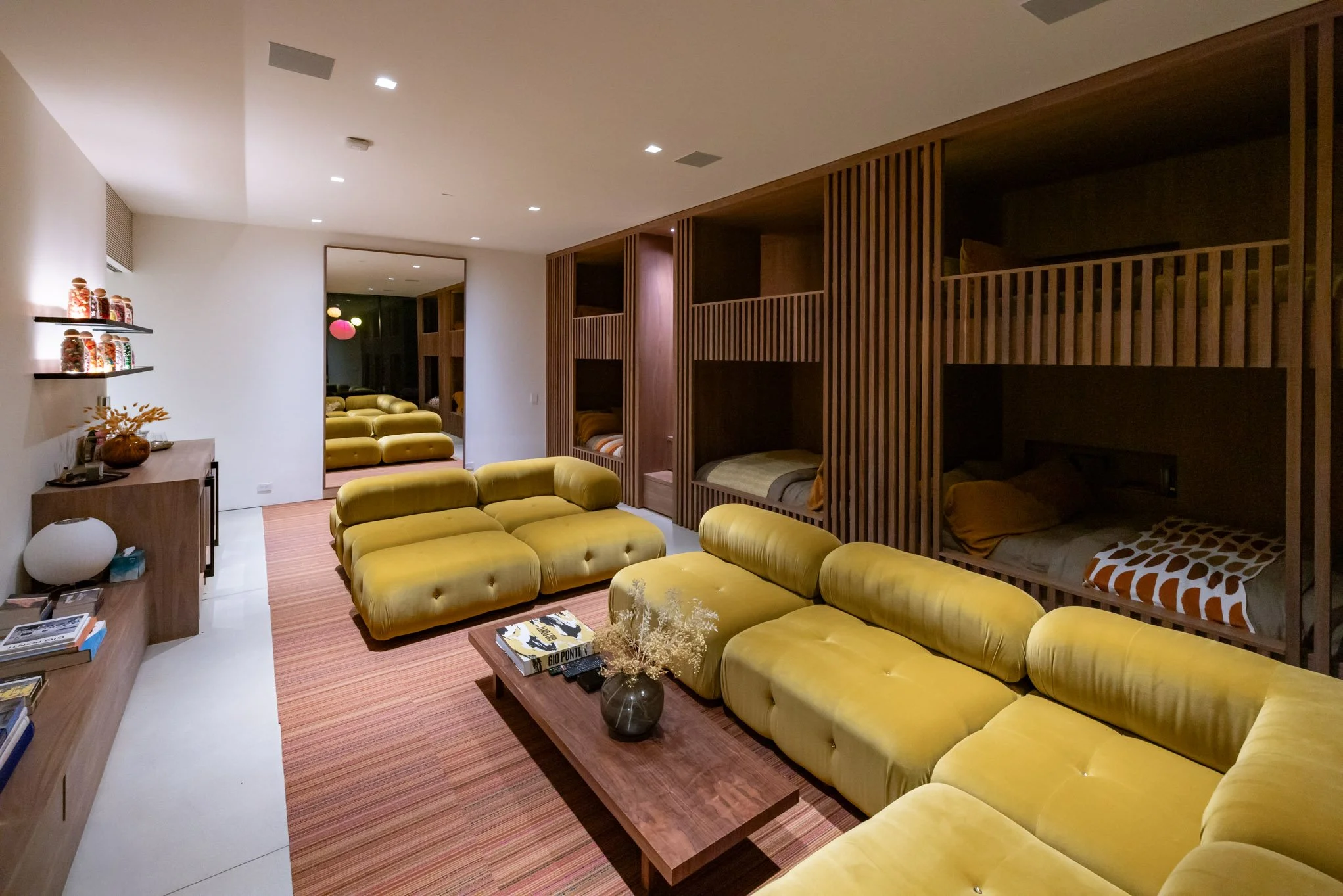 Modern living room with large yellow sectional sofa, wooden coffee table with books and a vase, built-in wooden sleeping pods with beds on elevated platforms, a mirror reflecting additional yellow seating, and wall-mounted shelves with decorative jar