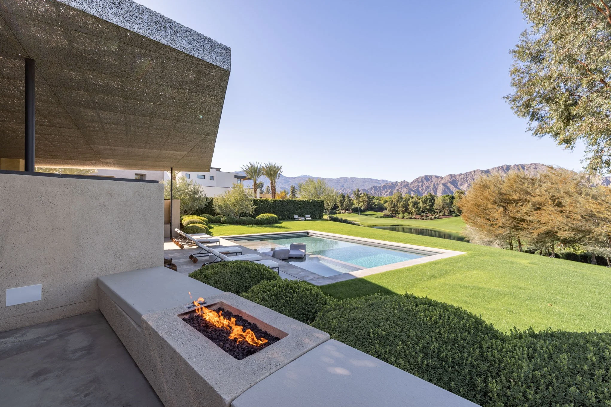 Modern house patio with built-in firepit, overlooking a lush backyard with a swimming pool, green lawn, trees, and distant mountains under a clear blue sky.