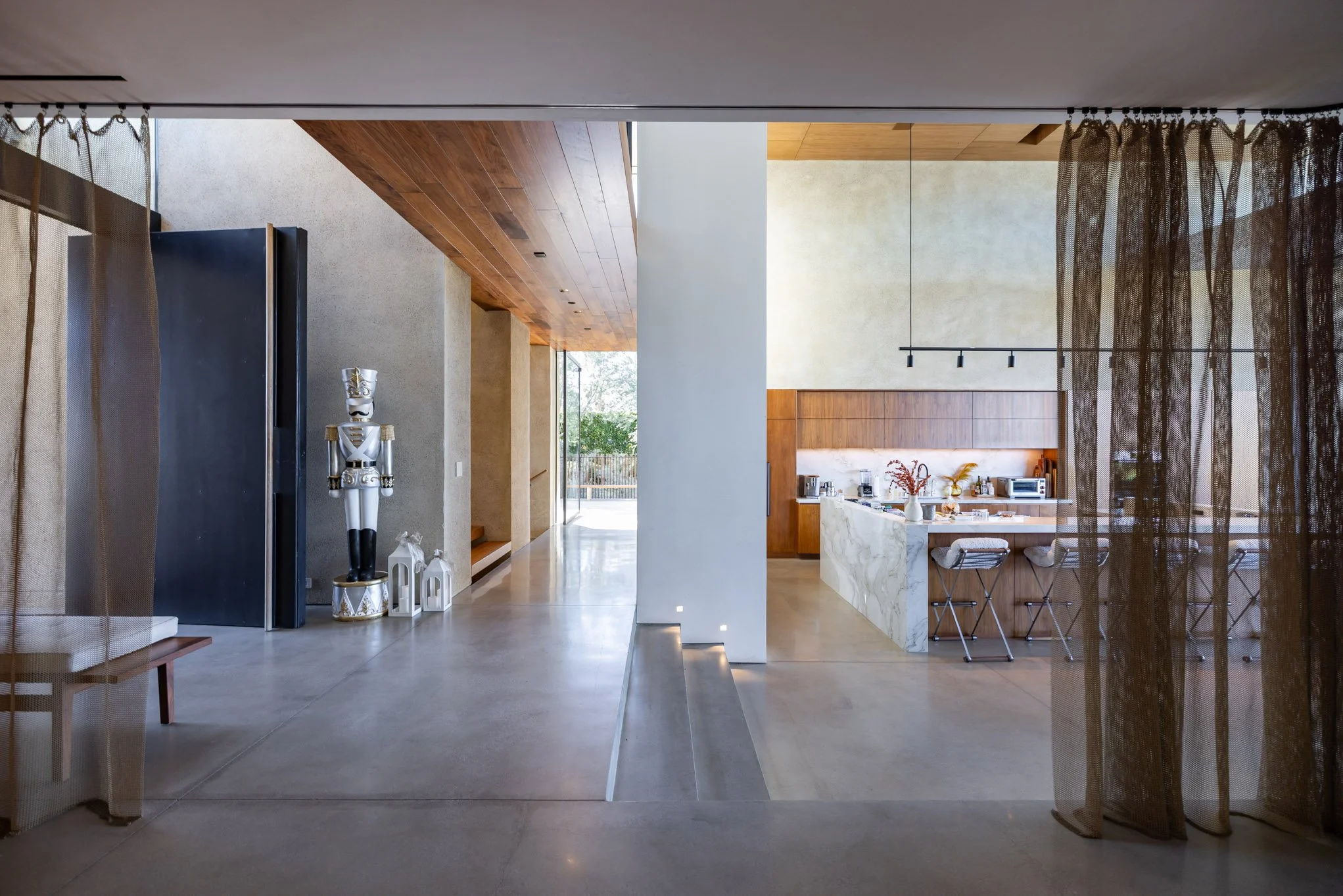 Modern open-concept kitchen and living area with wooden ceiling, large windows, and minimalist decor, including a nutcracker statue and lanterns near the entrance.