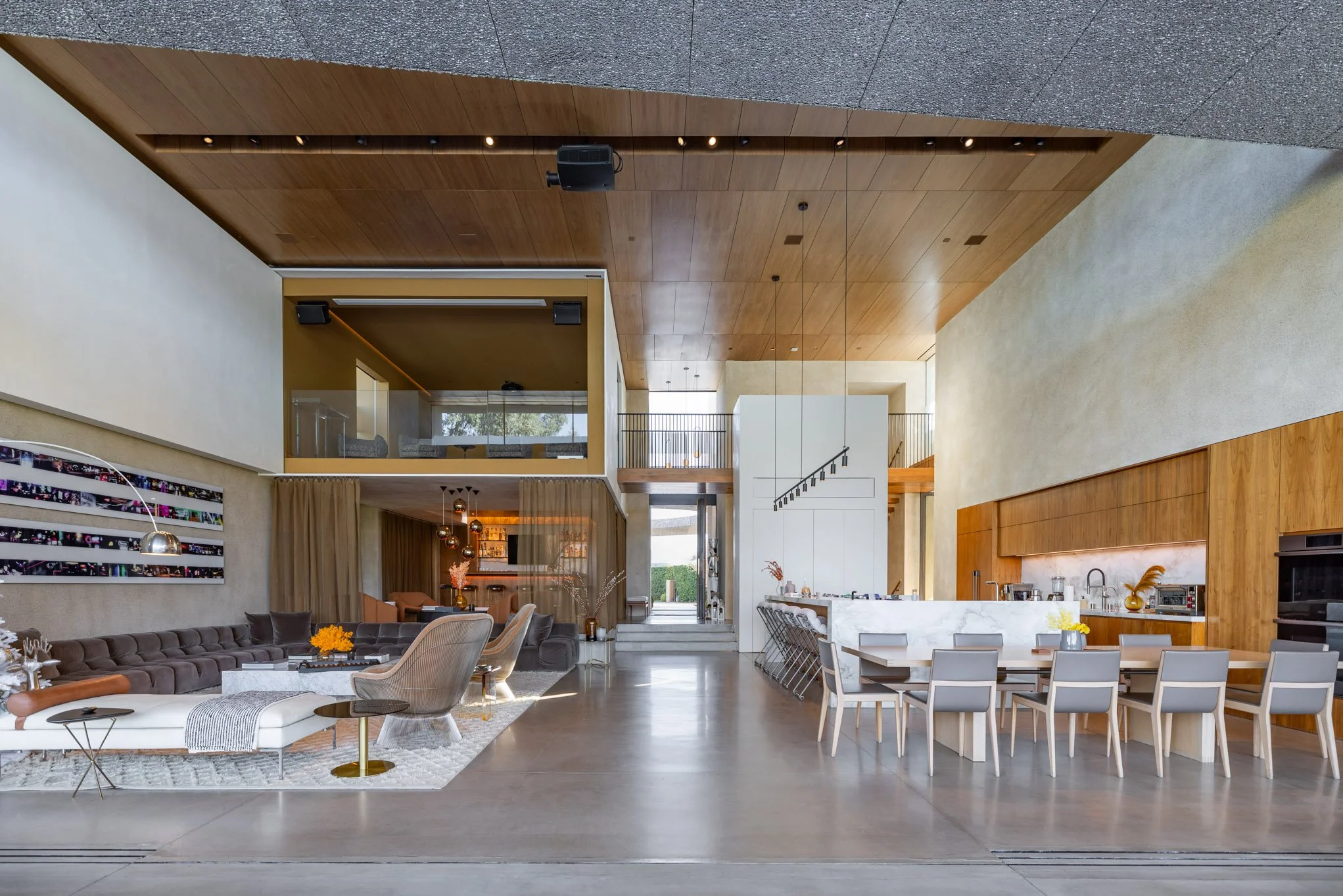 Modern open-concept living and dining area with high ceilings, wooden panel accents, and large windows, featuring a sectional sofa, armchairs, a dining table with chairs, and a kitchen island.