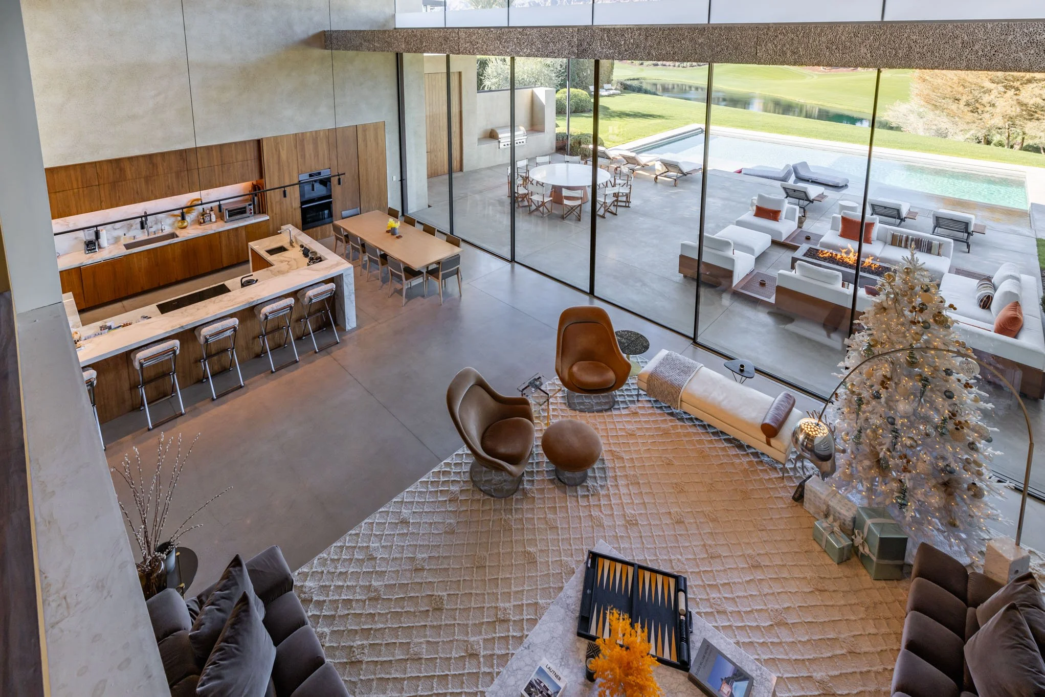 Interior of a modern living space with an open kitchen, dining, and seating areas. Large floor-to-ceiling windows reveal an outdoor patio with lounge chairs, a pool, and a lush green landscape. The room features a Christmas tree with presents underne