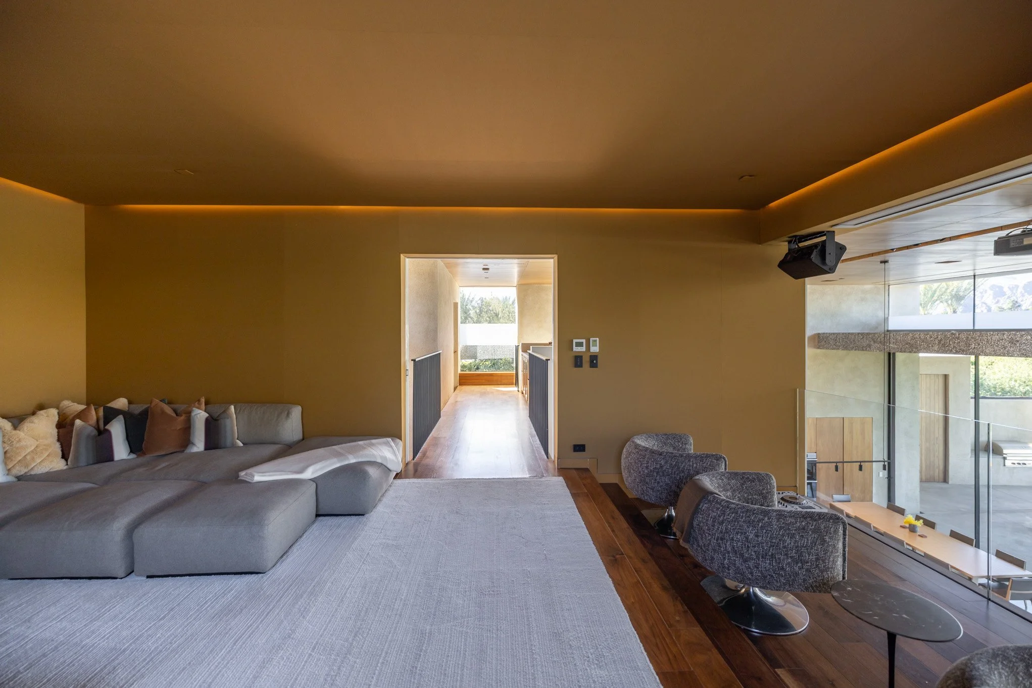 Modern living room with beige walls, large sofa with pillows, wooden floor, and view of outdoor patio through large windows.