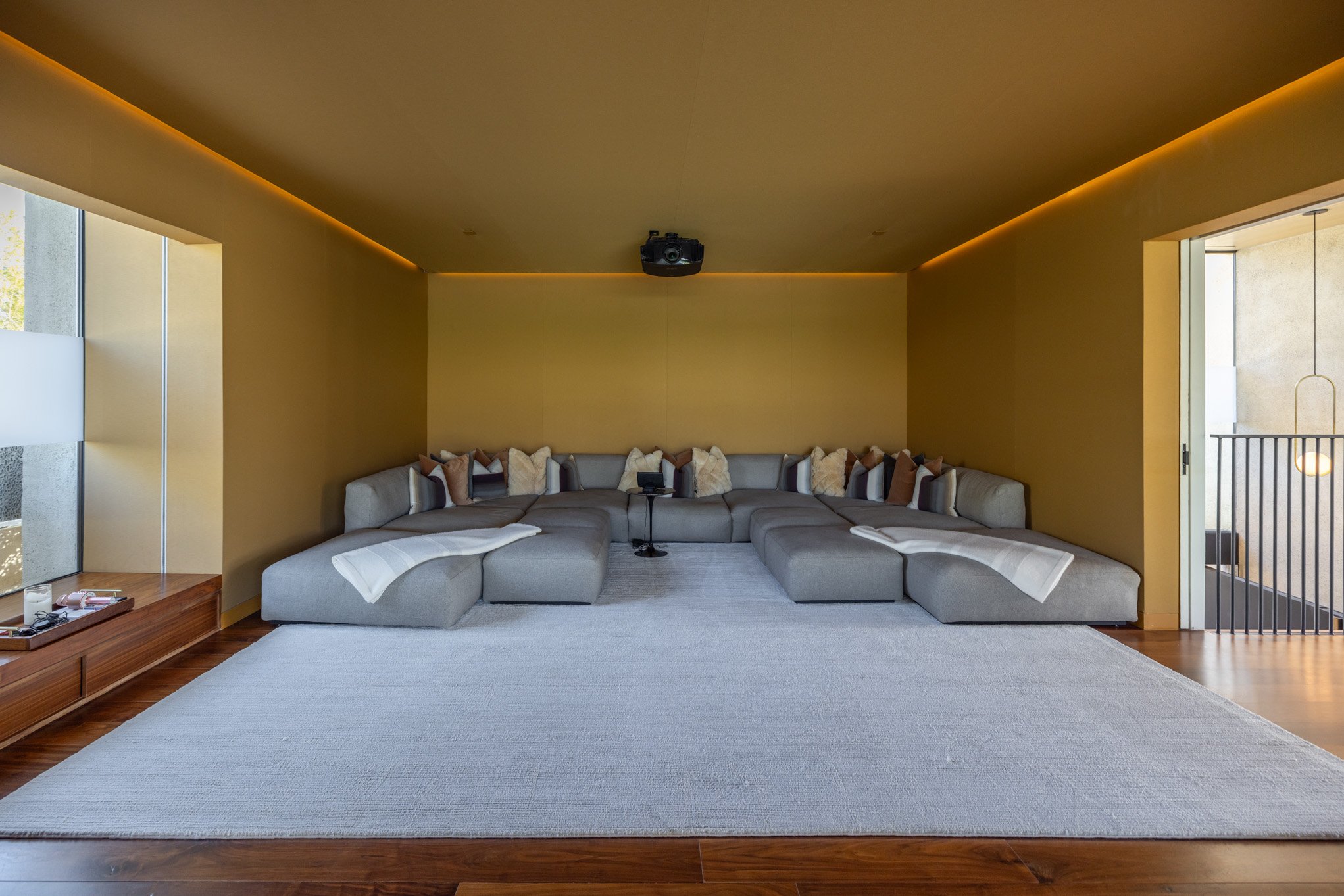 Modern living room with a large, light gray L-shaped sectional sofa decorated with various pillows, a white throw blanket, and a black overhead projector mounted on the ceiling. Windows on the left side allow natural light and a view of greenery, wit