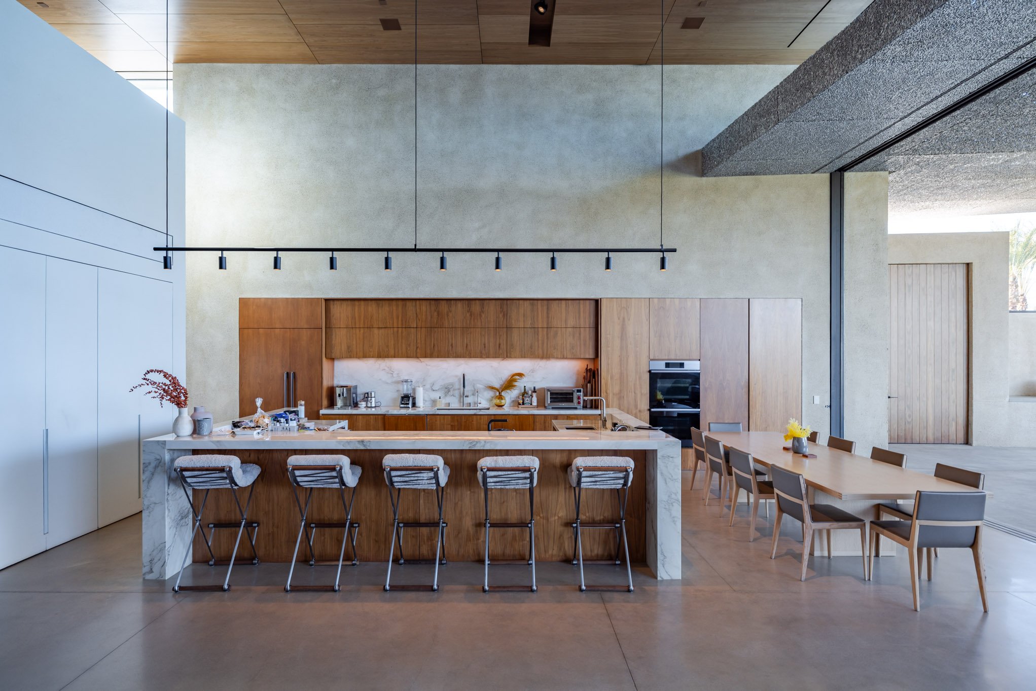 Modern kitchen with wooden cabinets, marble kitchen island with barstools, and a dining table with chairs. Light-colored walls and ceiling, with large windows letting in natural light.
