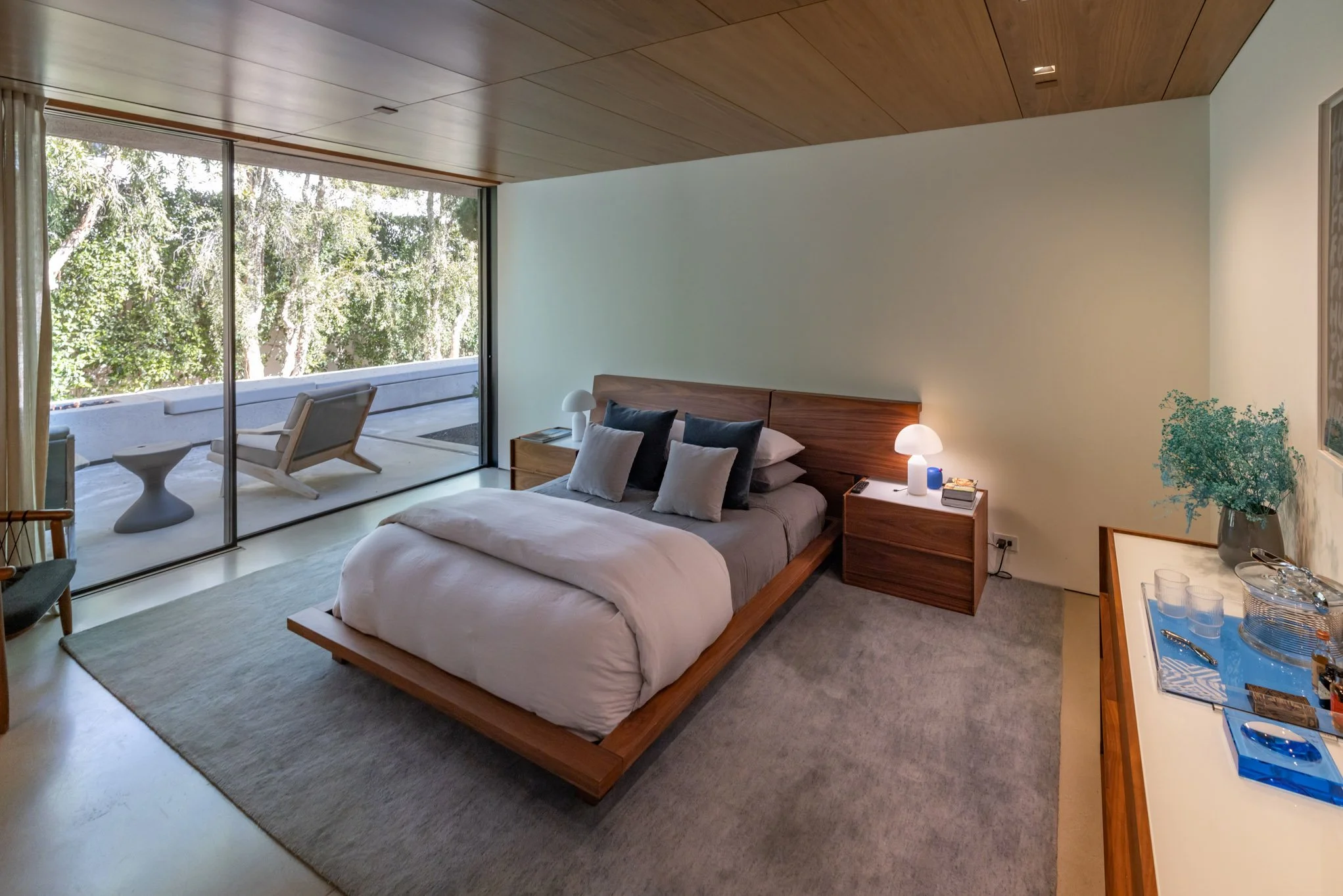 A bedroom with a large glass sliding door leading to a balcony with outdoor seating. The room has a bed with pillows and a wooden headboard, two nightstands with lamps, a dresser with a plant and glassware, and neutral-colored walls and carpet.