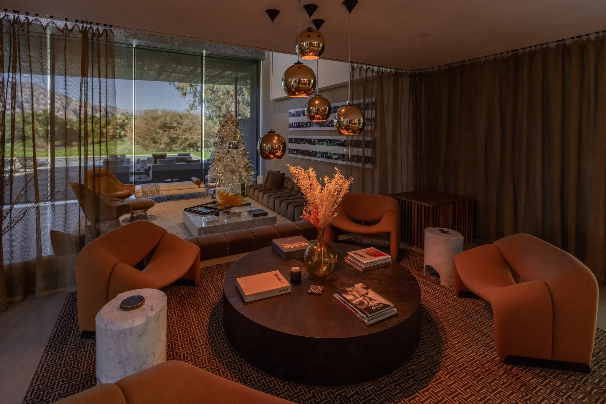 Modern living room with brown and beige furniture, a round coffee table with books, a decorative vase with dried flowers, and pendant lights. There's a Christmas tree in the background near large glass windows showing an outdoor patio and green lands