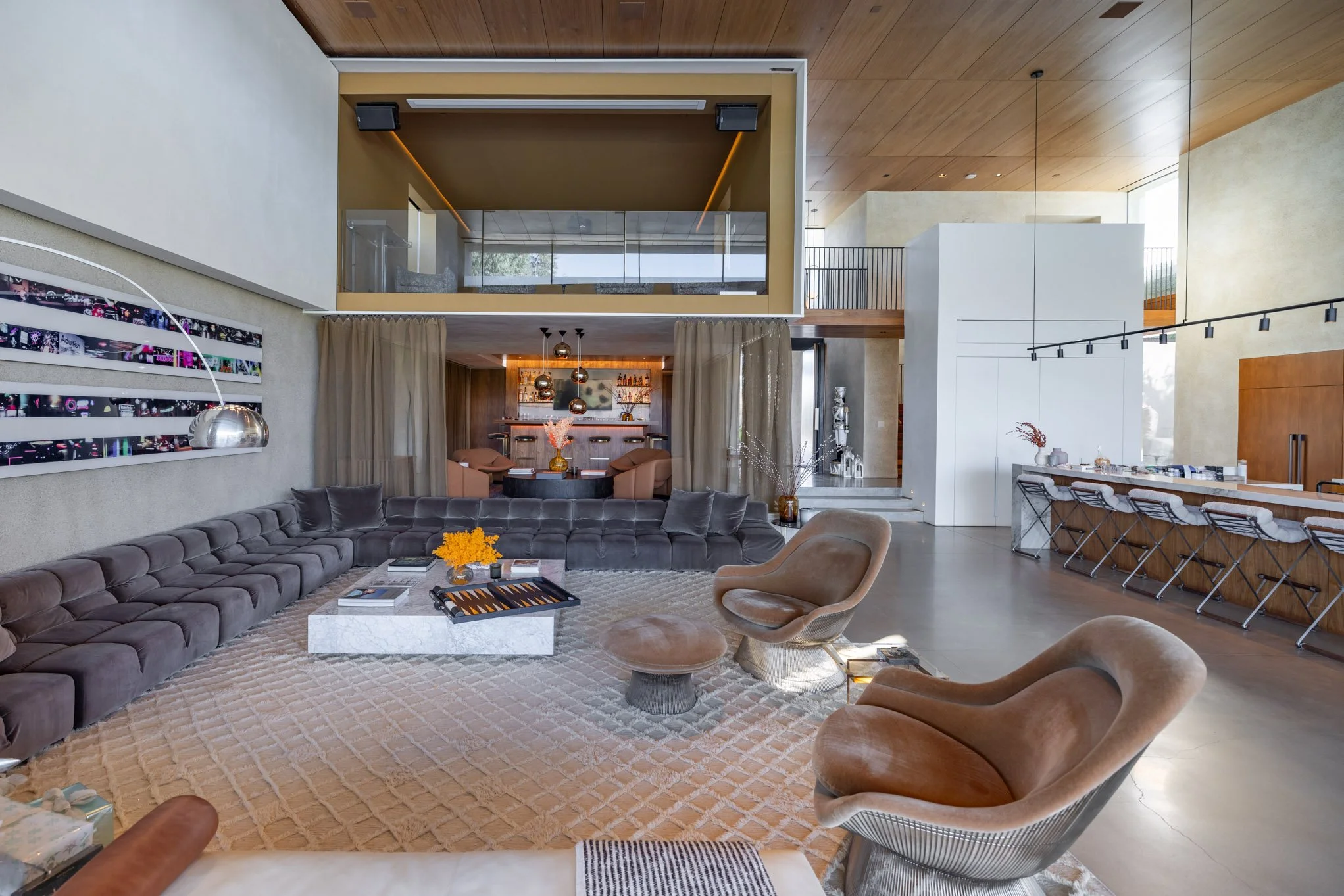 A spacious, modern living room with a large, gray sectional sofa, two beige armchairs, and a center coffee table on a patterned rug. There is a bar area with barstools on the right, a dining area with chairs in the background, and a second-floor loft