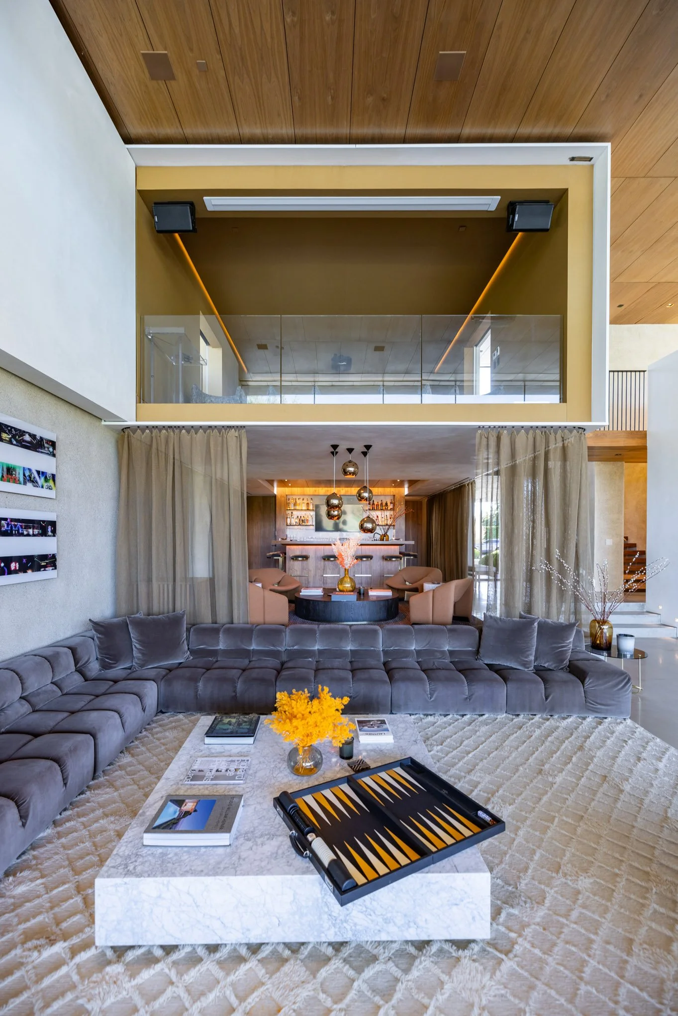 Modern living room with a large gray sectional sofa, a white marble coffee table, and decorative items including books, magazines, a yellow flower arrangement, and a backgammon game. The background features a bar area with peach-colored chairs, penda