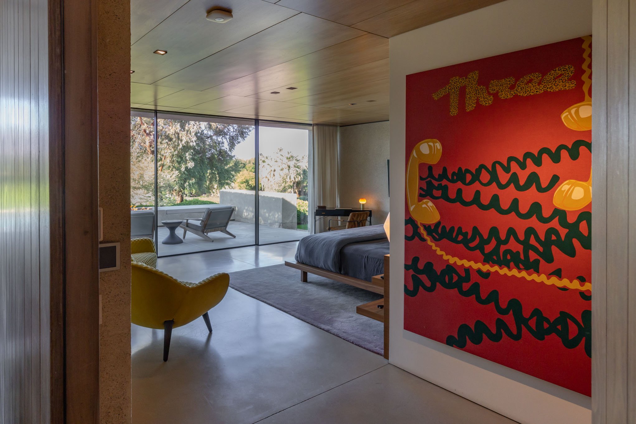 Modern bedroom with a large sliding glass door leading to an outdoor patio, a yellow armchair near a wall, a bed with a dark duvet, a desk with a lamp, and abstract art with red, yellow, and black colors on the wall.