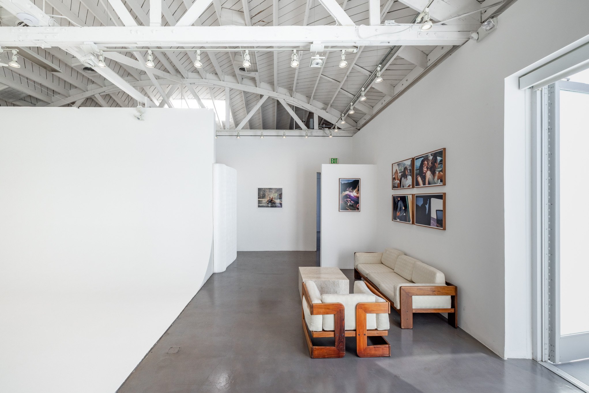 Interior view of an art gallery with white walls, featuring framed photographs and artwork. The space has a high, white-painted industrial ceiling with exposed beams and track lighting. There is modern white and wooden seating in the corner near a la