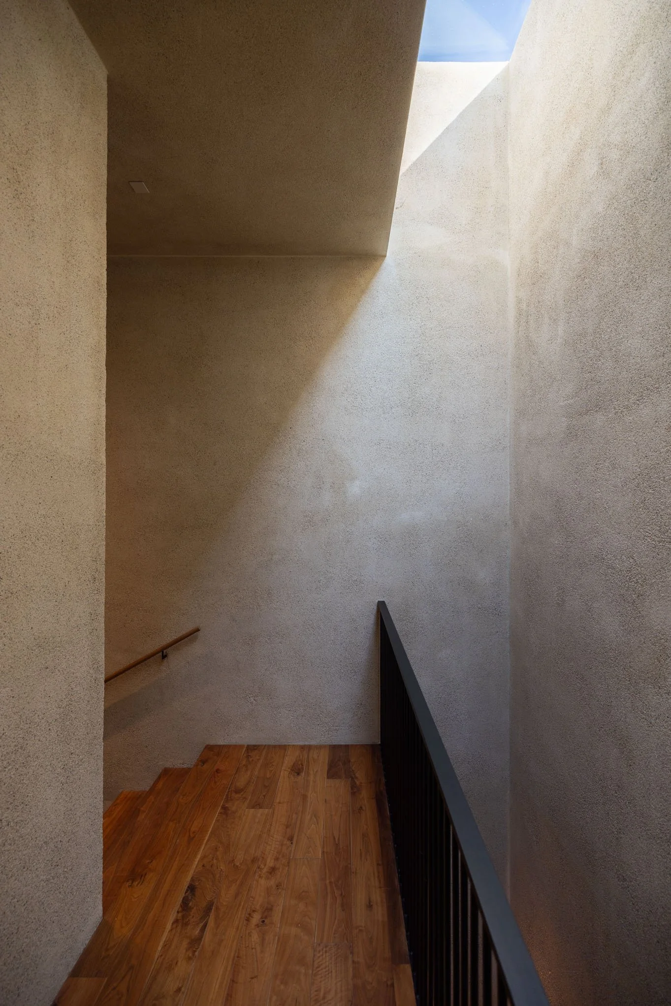 Interior view of a staircase landing with textured beige walls, a wooden floor, a black railing on the right, and a skylight allowing sunlight to illuminate the space.