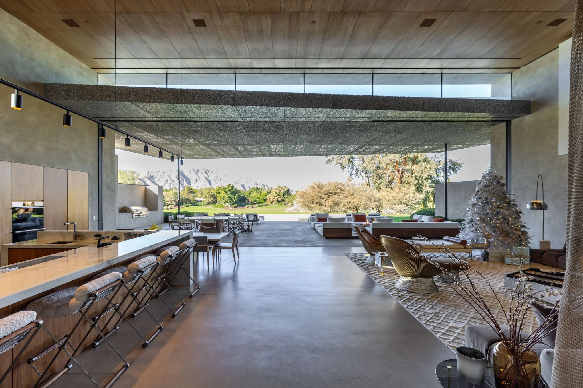 Modern open-concept living space with kitchen island, bar stools, outdoor seating, and a decorated Christmas tree, overlooking a green landscape.