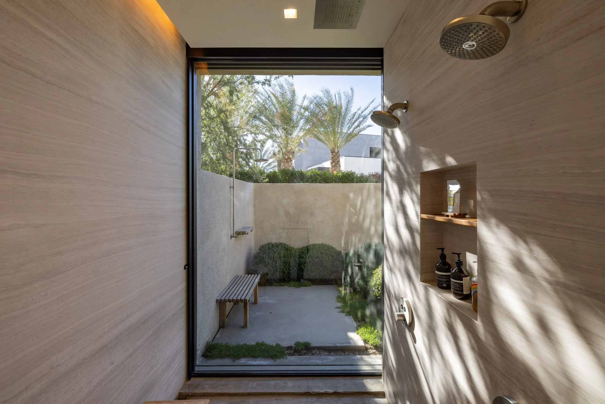 Modern outdoor shower area with a large glass window, palm trees visible outside, and built-in shelves with toiletries.