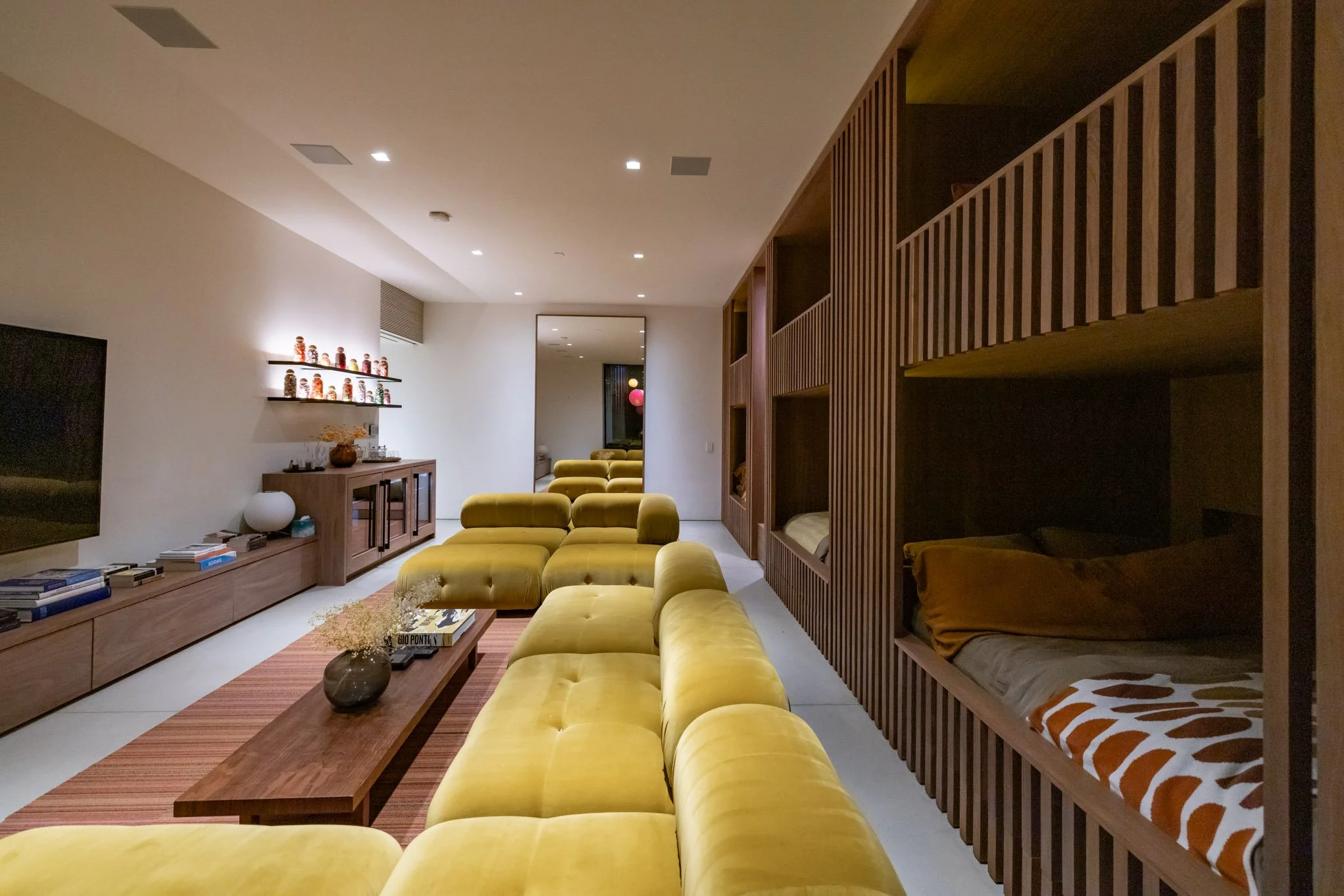 Modern living room with yellow velvet sectional sofa, wooden coffee table, wall-mounted shelves with decorative jars, and built-in beds on wooden partitions.