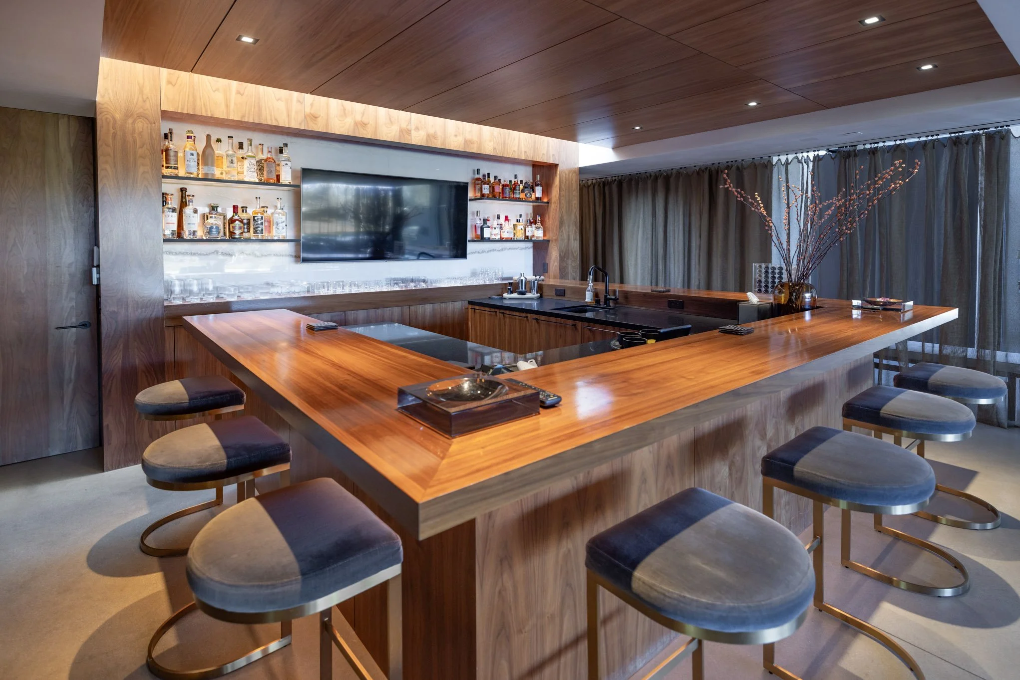 Modern bar with a wooden U-shaped counter, six barstools with velvet seats, a large TV, bottles of alcohol on shelves, a vase with branches, and a curtain-covered window.