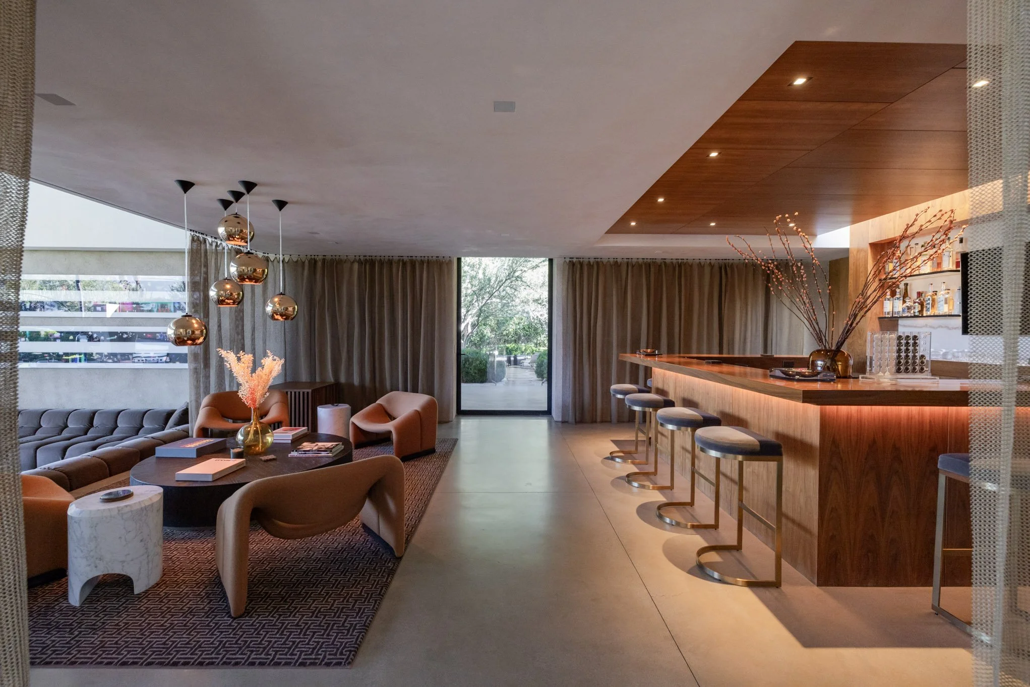 Modern lounge with a seating area, round table, armchairs, sofa, and pendant lights, adjacent to a bar with barstools, a vase with branches, and a view of the outdoors through large glass doors.